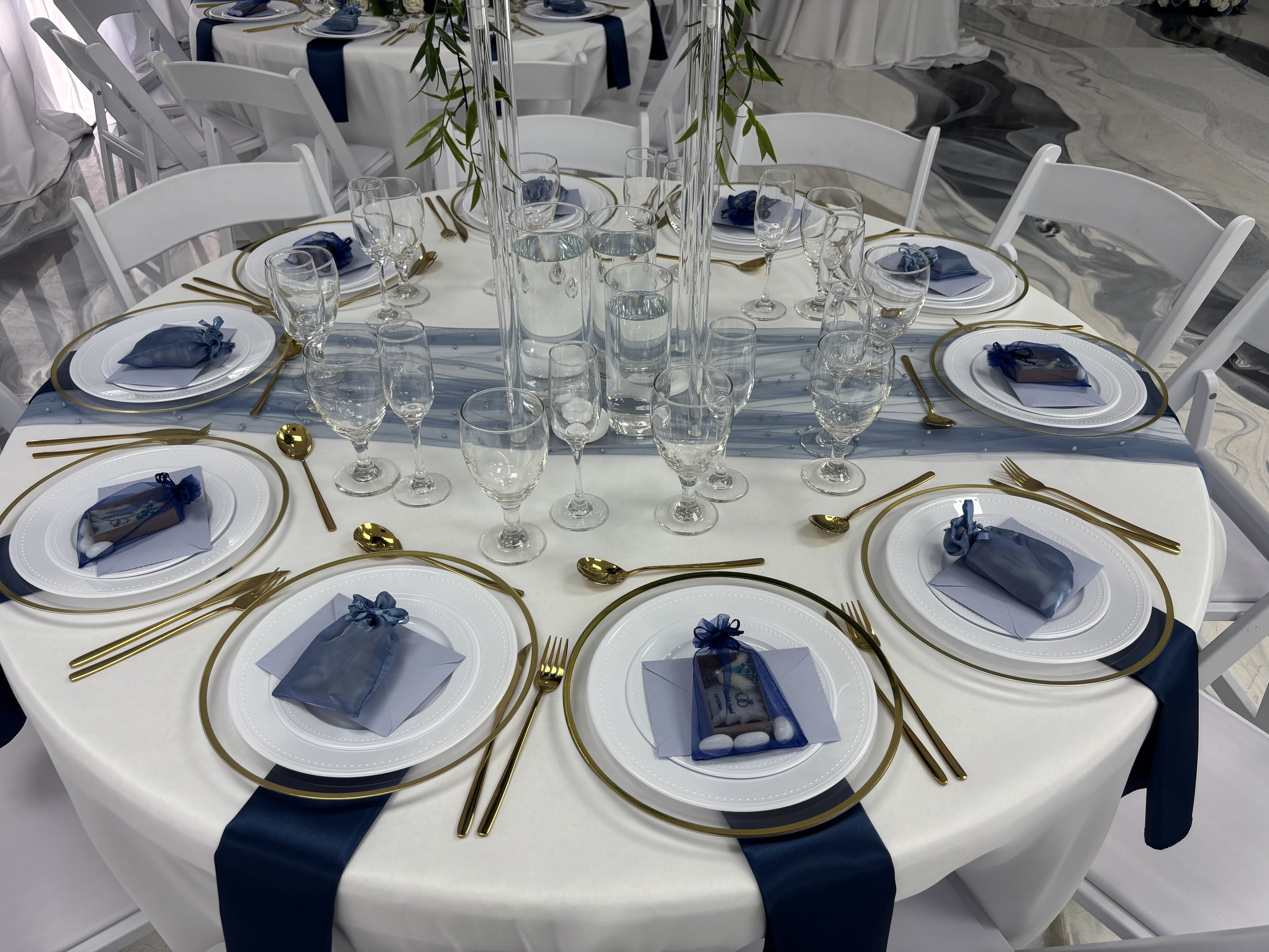 Round table set for a formal event with white tablecloth, navy blue sashes, and gold cutlery. Each place setting includes a white plate, a small wrapped favor with a blue ribbon, and a folded napkin. Clear glasses are arranged around the table, and a