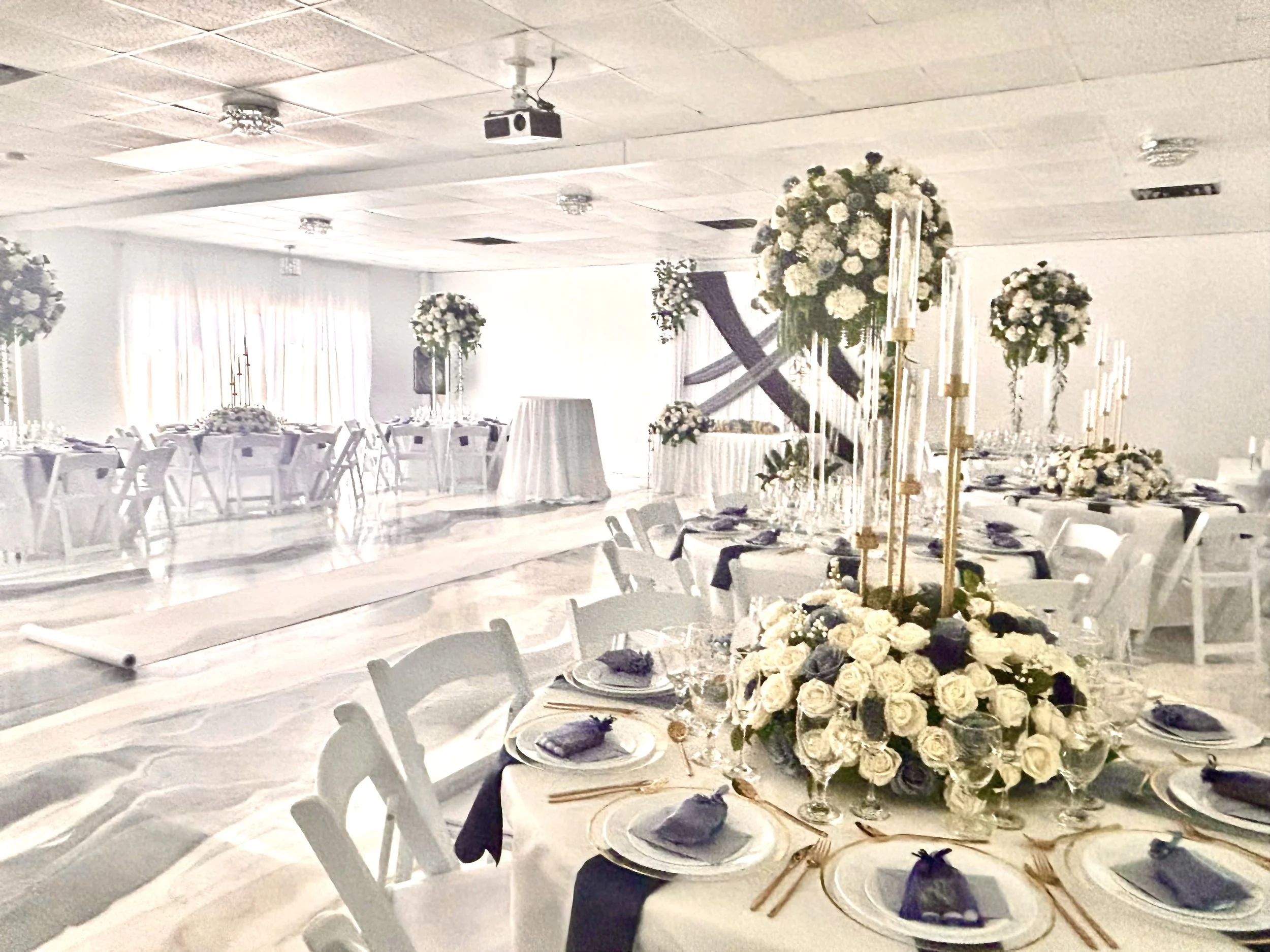 Elegant banquet hall decorated for a wedding reception with white floral centerpieces, gold accents, and purple napkins on white tables.