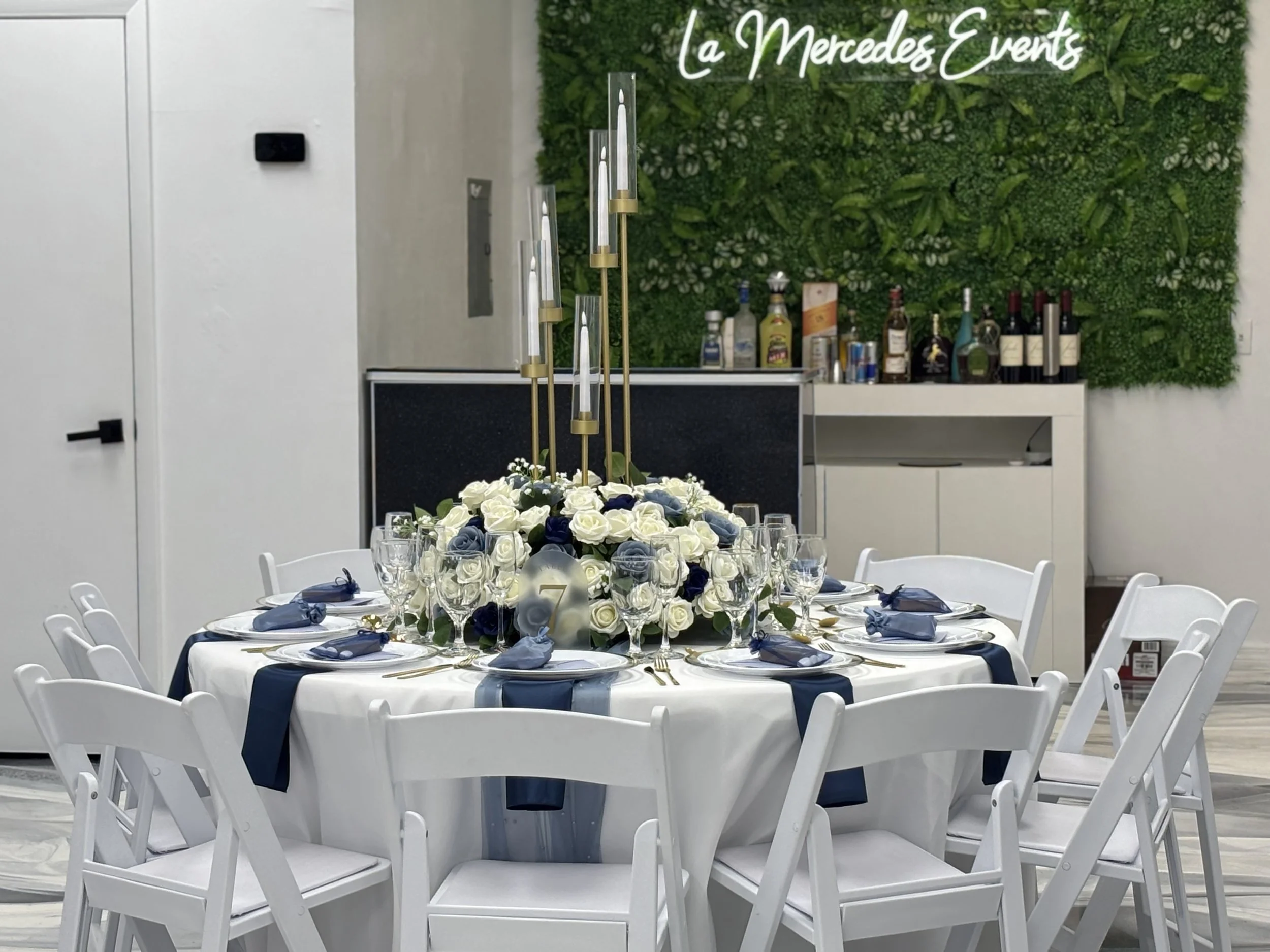 Round dining table decorated for a formal event with white and navy blue flowers, candles, and glassware, set for eight people, with a backdrop of a green wall and sign reading 'La Mercedes Events'.