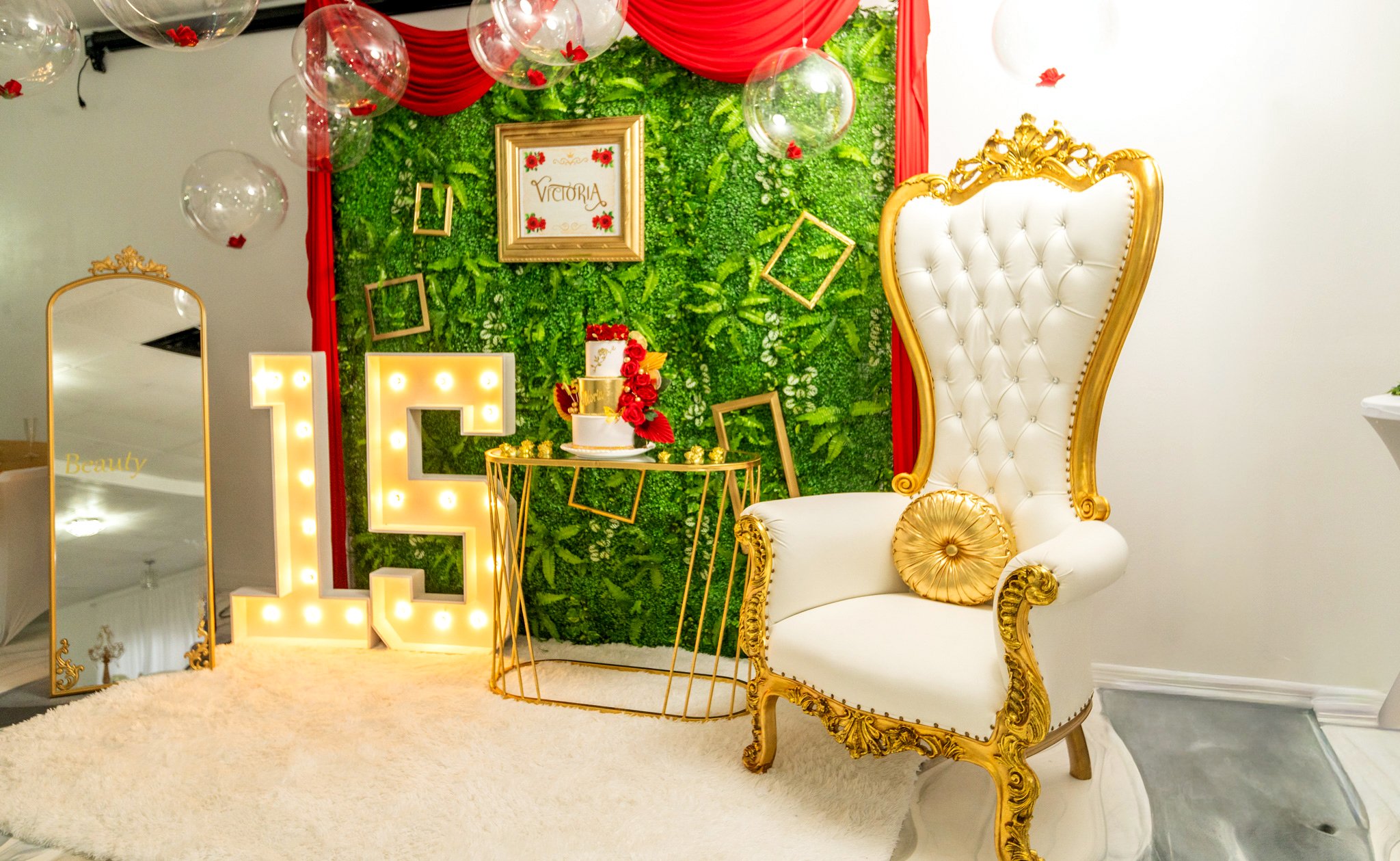 Elegant party setup with a large illuminated letter E, a white and gold ornate high-back chair, a green leafy backdrop with a framed sign reading "Victoria," floating glass balloons with red accents, and a white fluffy rug.