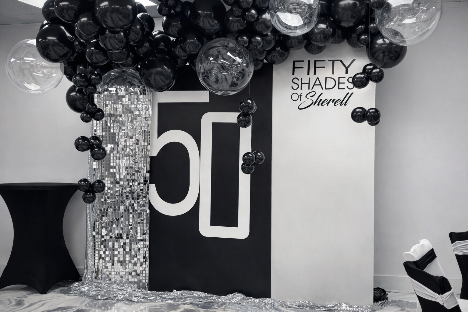 Decorative backdrop with black and silver balloons, a black panel with Marilyn Monroe's face outline, and a sign that reads "FIFTY SHADES of Sherlock", in a monochrome black, silver, and white color scheme, likely for a themed event or party.