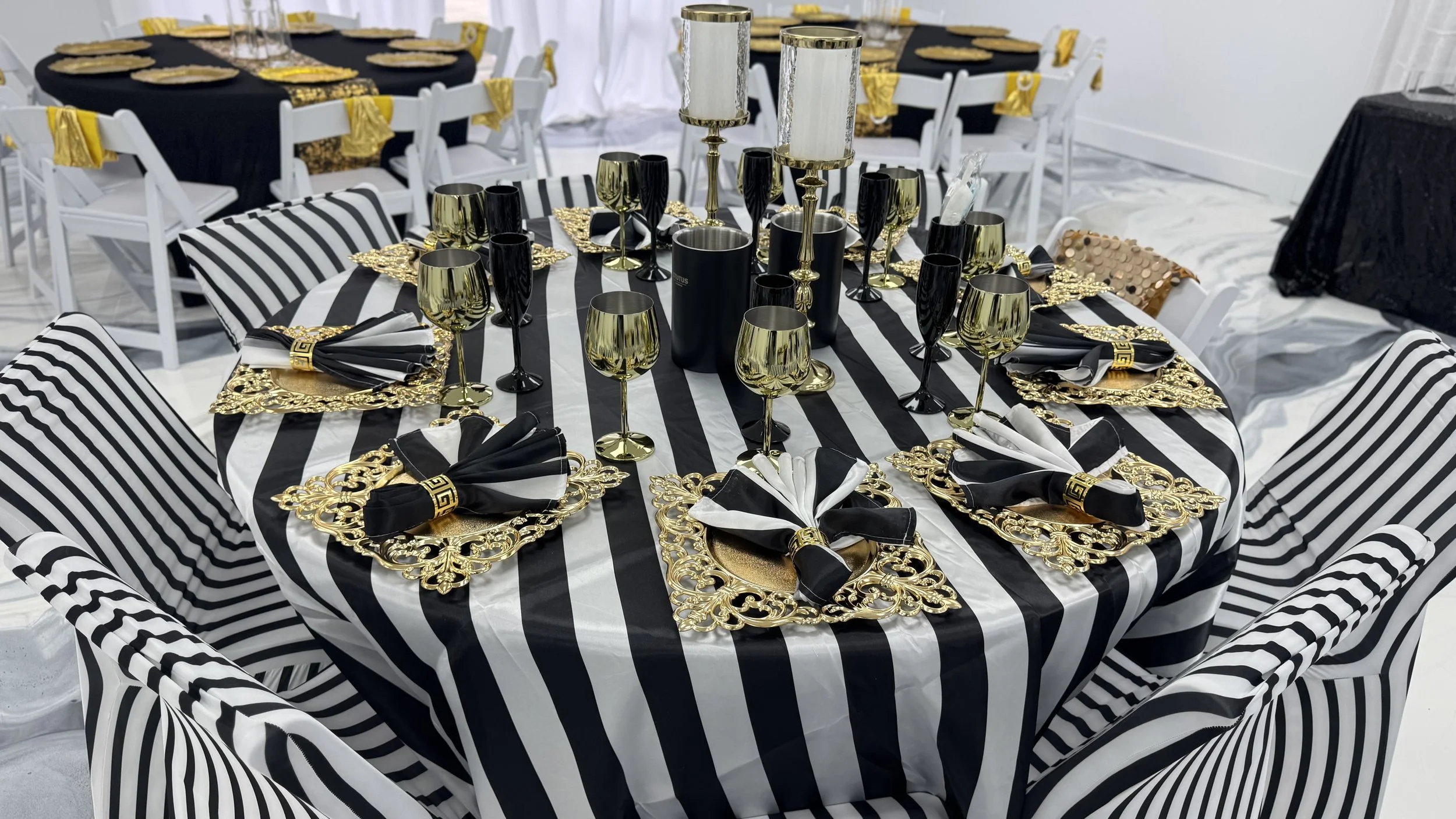 A round banquet table decorated with black and white striped tablecloth and matching striped chairs. The table setting features gold ornate placemats, black and white napkins with gold napkin rings, and gold and black glasses. In the center, there ar