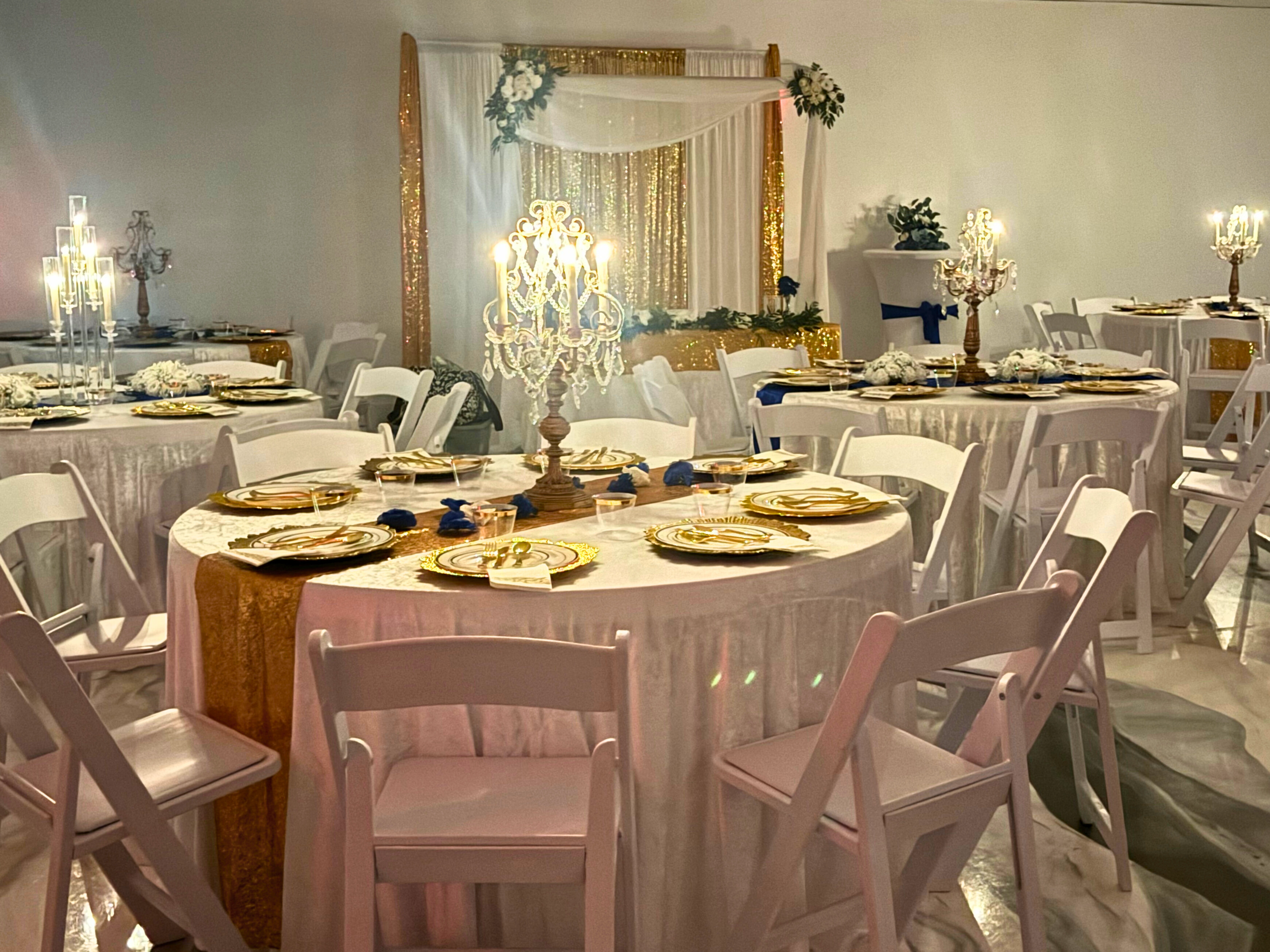 Elegant event space with round tables decorated with gold-rimmed plates, white and navy blue napkins, floral centerpieces, and candelabras, set for a celebration or wedding reception.