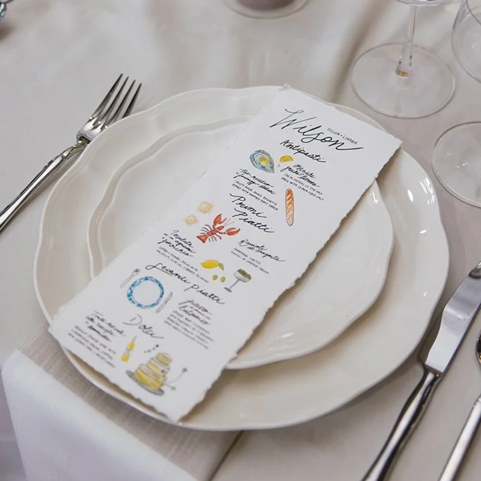 A dinner table setting with a white plate, a fork on the left, a knife on the right, and several empty wine glasses. There is a torn-edge paper menu placed on top of the plate with colorful illustrations and handwritten-style text.