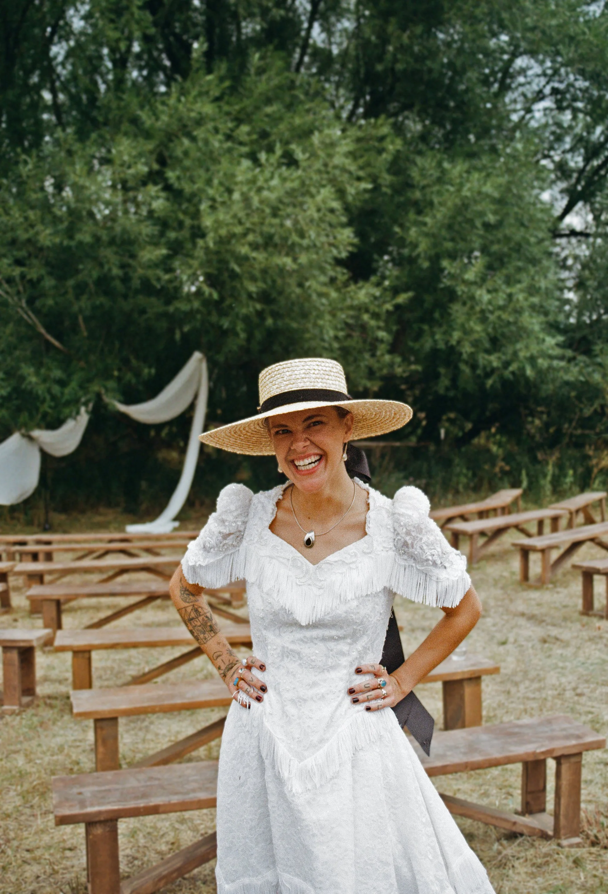 Woman in white dress and large straw hat smiling outdoors, with benches and green trees in the background.