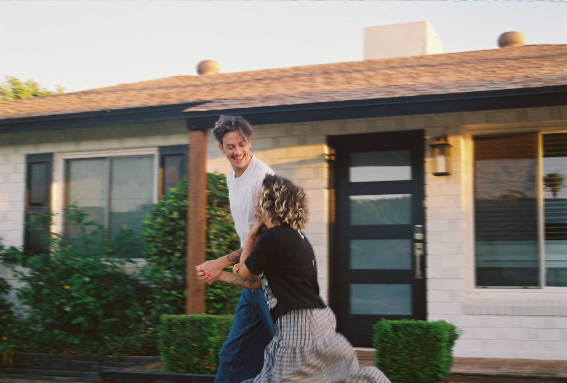A man and a woman are playing outside in front of a house, holding hands and smiling at each other during daytime.