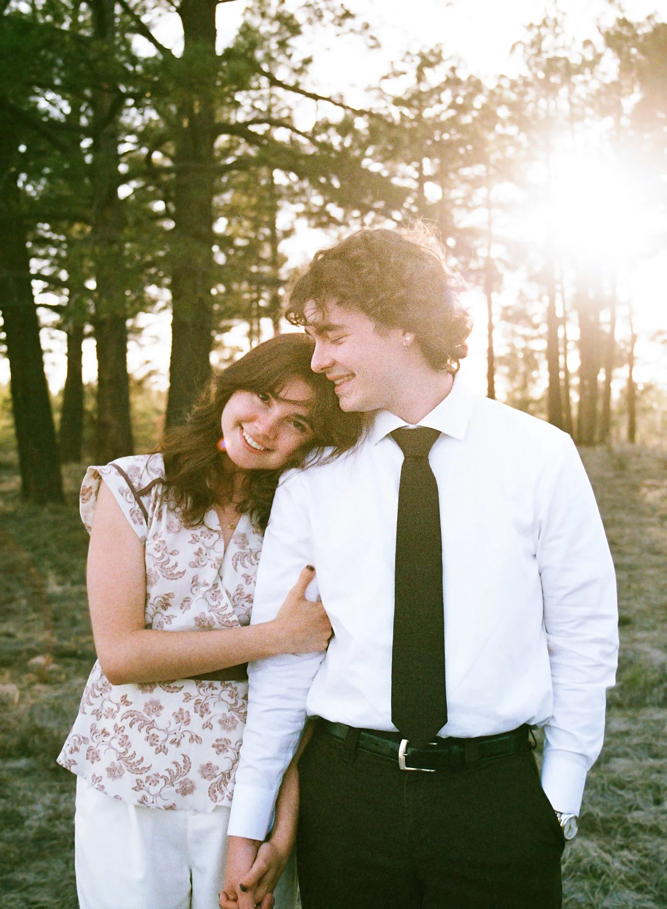 A smiling woman and man holding hands, leaning on each other, outdoors in a forest during sunset.