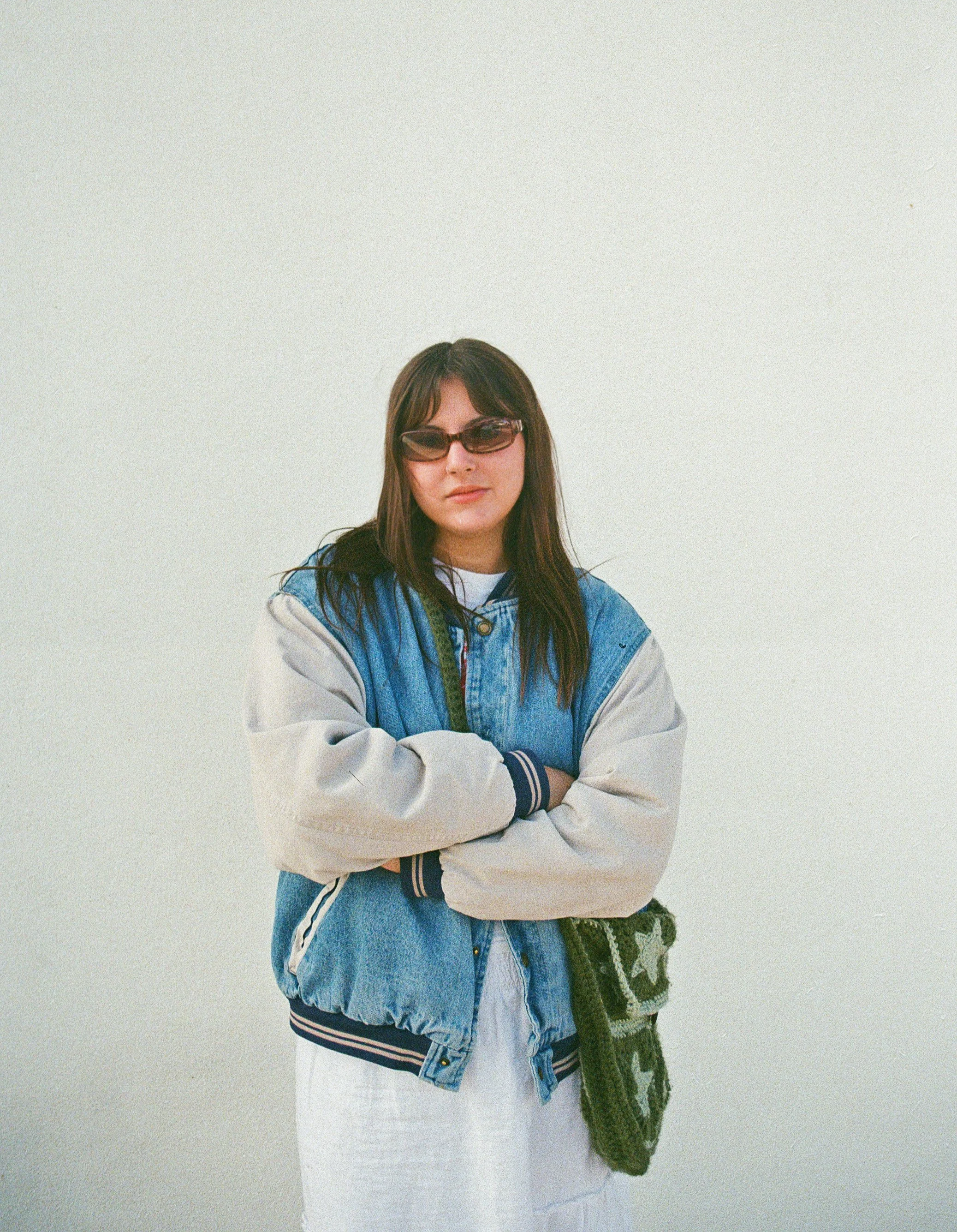 Young woman with long brown hair wearing sunglasses, a light blue and white varsity jacket, and a white dress, standing against a plain white wall.