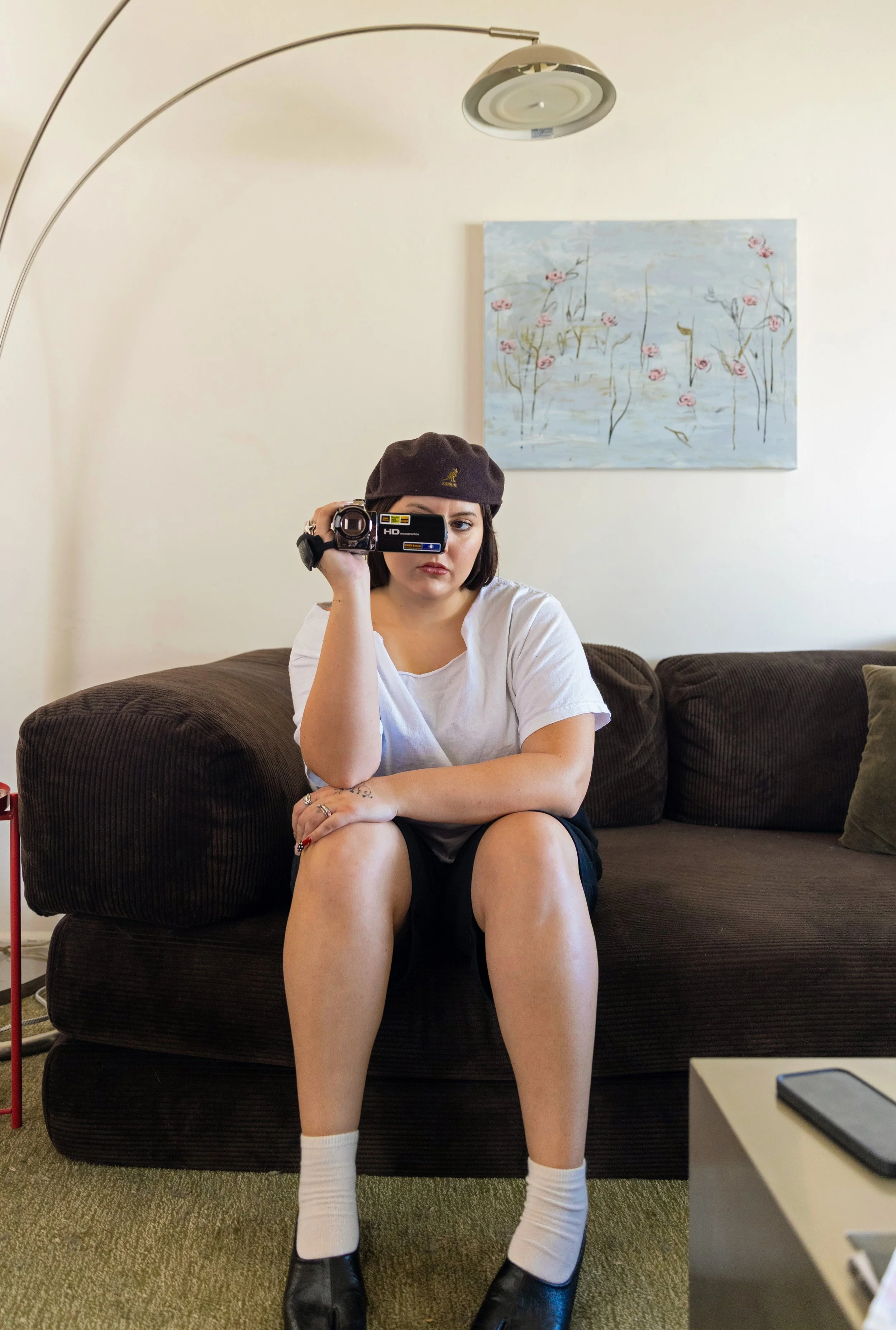 A woman sitting on a brown couch, wearing a white t-shirt, black shorts, white socks, and black shoes, taking a photo with a vintage video camera. She has a brown beret and short dark hair. There is a colorful abstract painting on the wall behind her and a modern floor lamp above.