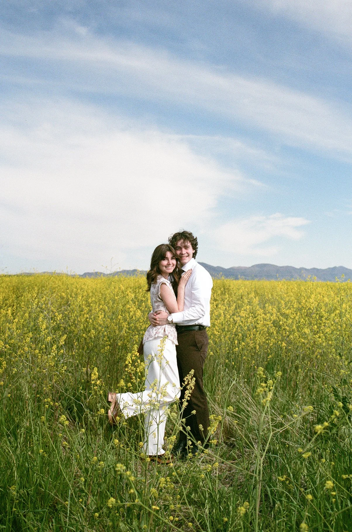 A happy couple embracing in a yellow flower field under a blue sky with clouds.