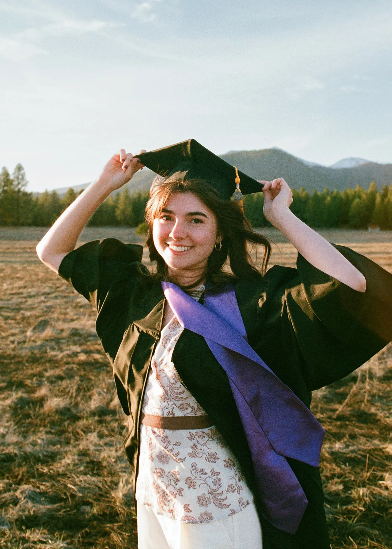 A young woman wearing a graduation gown and cap standing outdoors during sunset, smiling, with mountains and trees in the background.