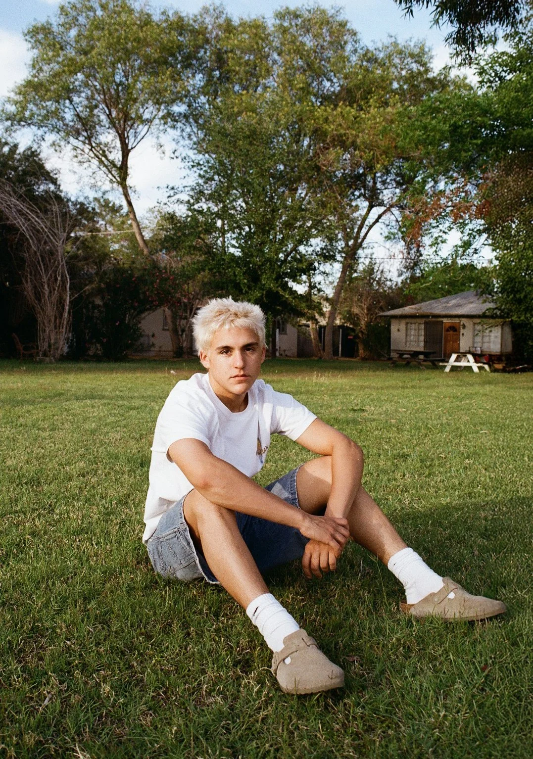Young person with bleached blonde hair sitting cross-legged on the grass in a backyard, with trees and a small house in the background.