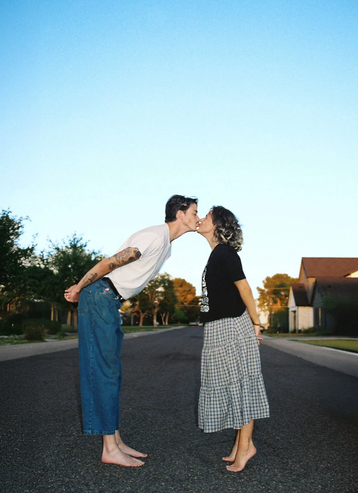 A man and woman are kissing each other on the lips while standing barefoot on an empty street at sunset, with houses and trees in the background.