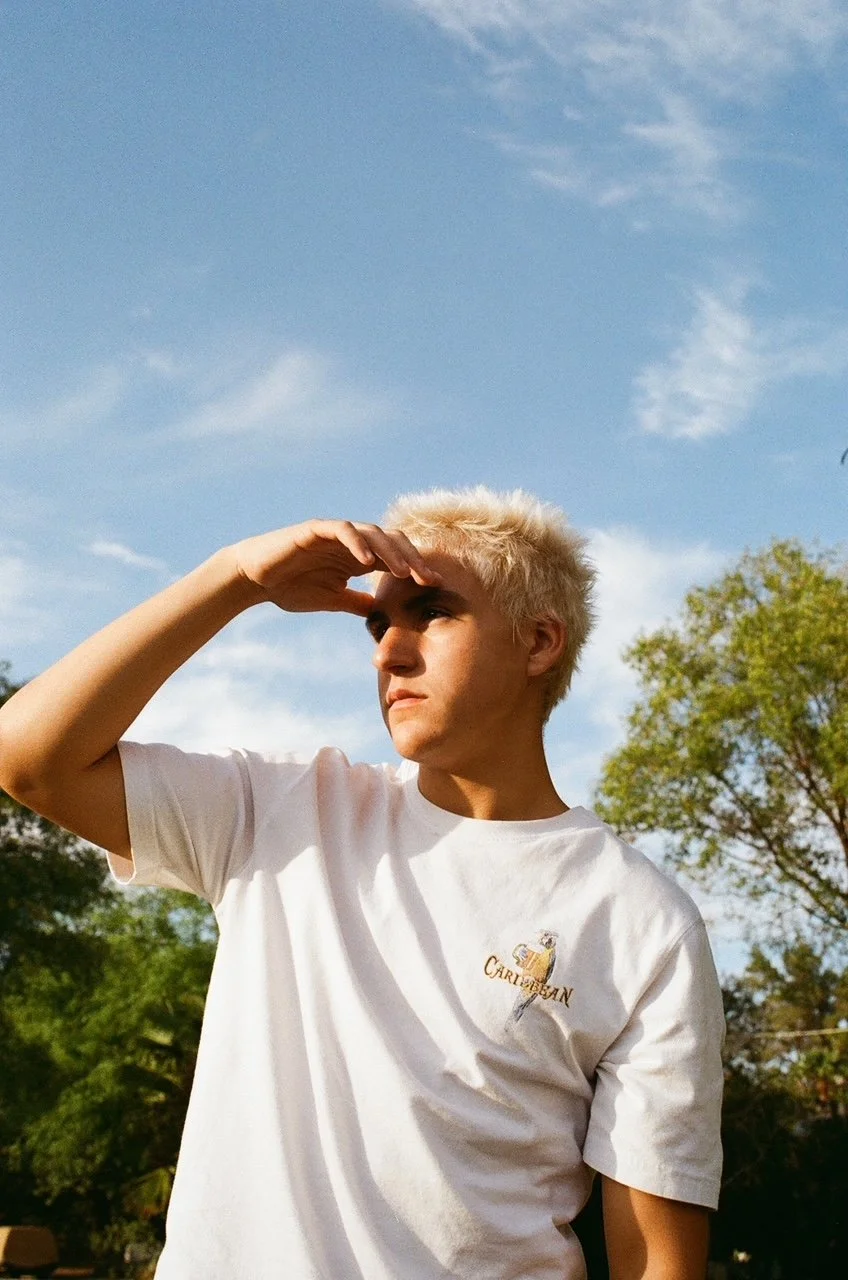 Young man with blonde hair wearing a white t-shirt standing outdoors, looking into the distance with a hand shielding his eyes from the sun, against a backdrop of blue sky and green trees.