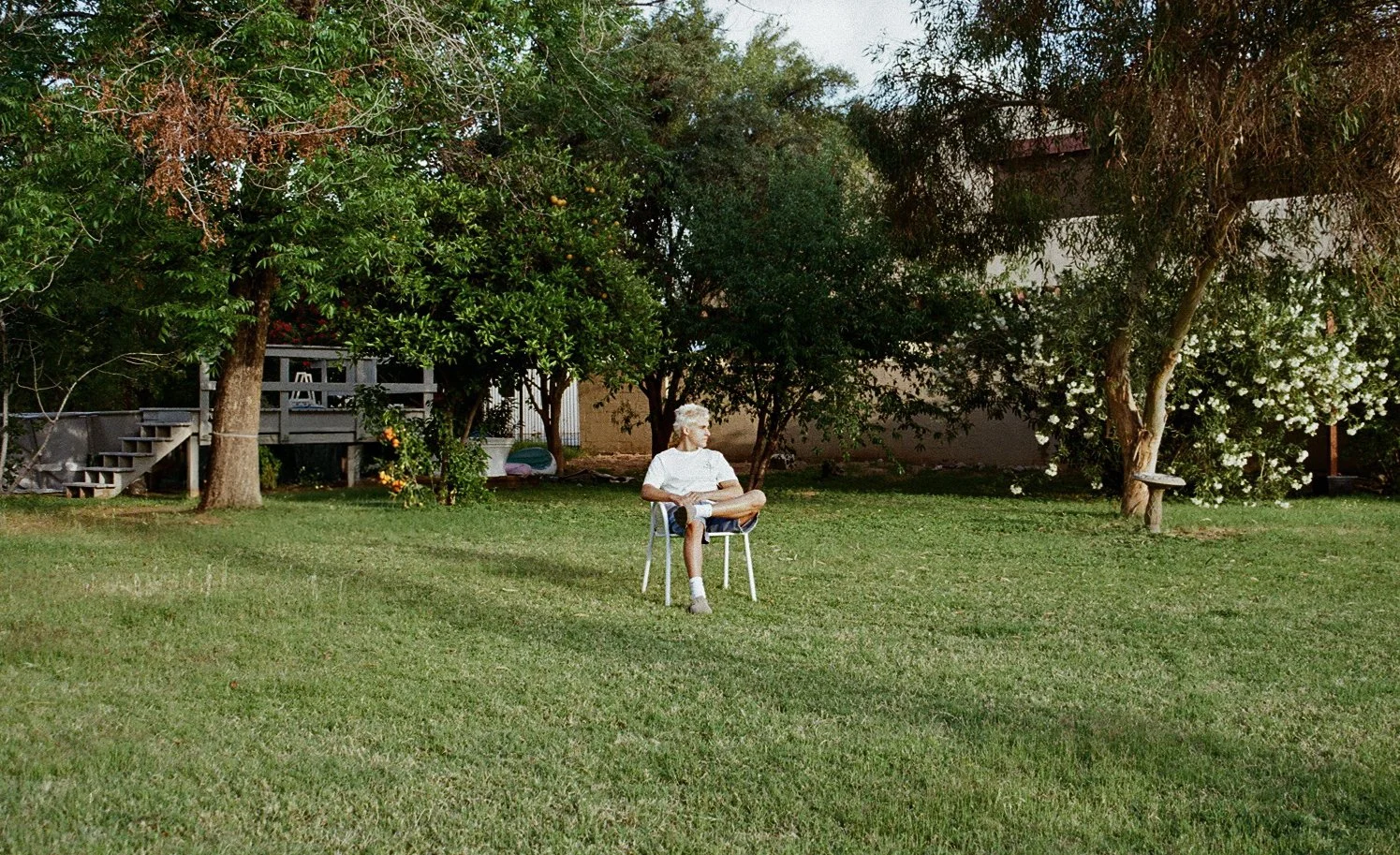A person with grey hair sitting cross-legged on a chair in a backyard with green grass, surrounded by trees and bushes, some with white flowers. There is a wooden staircase and a house in the background.