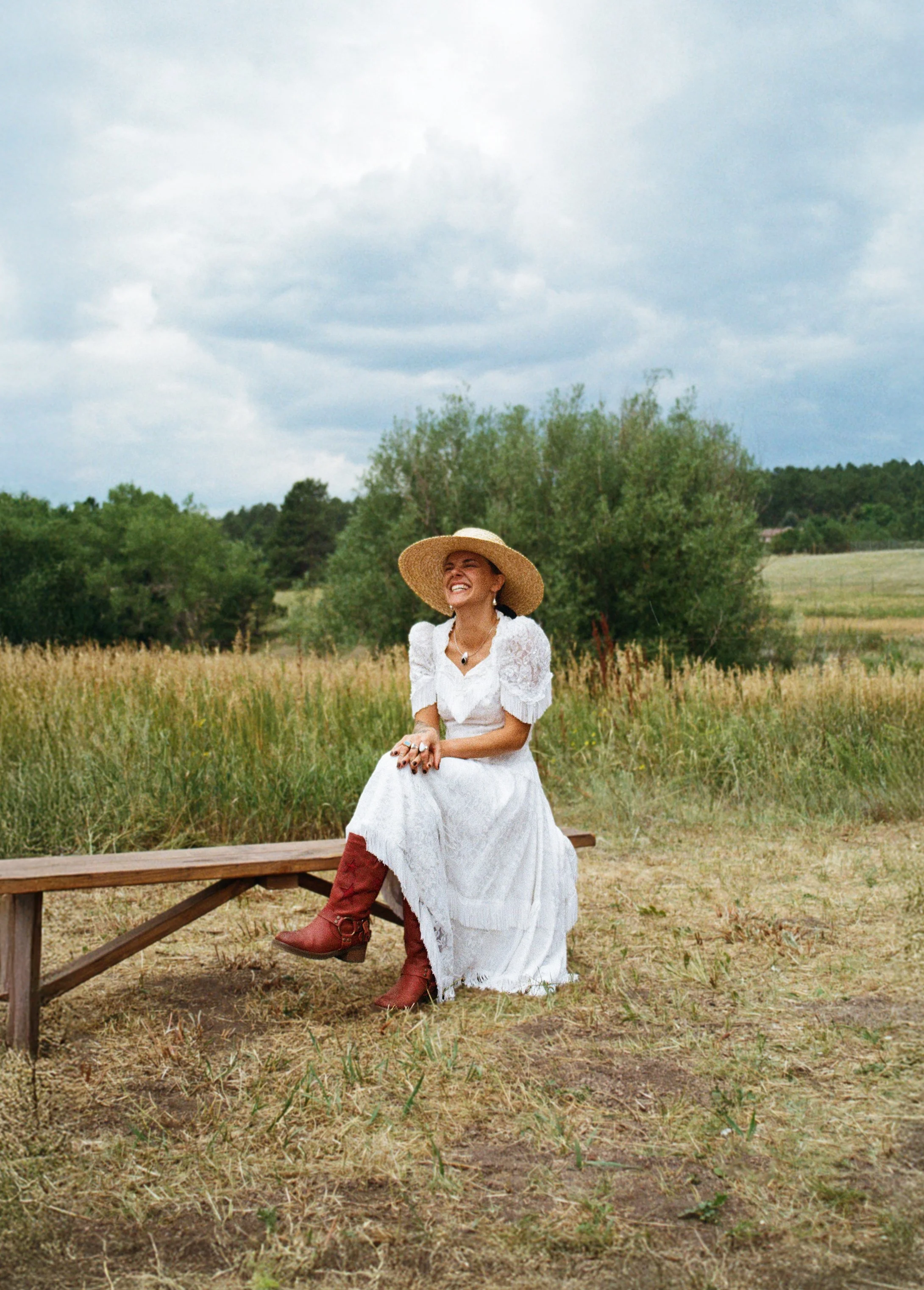 A woman in a white dress and wide-brimmed straw hat sitting on a wooden bench outdoors, smiling with a background of green trees and a cloudy sky.