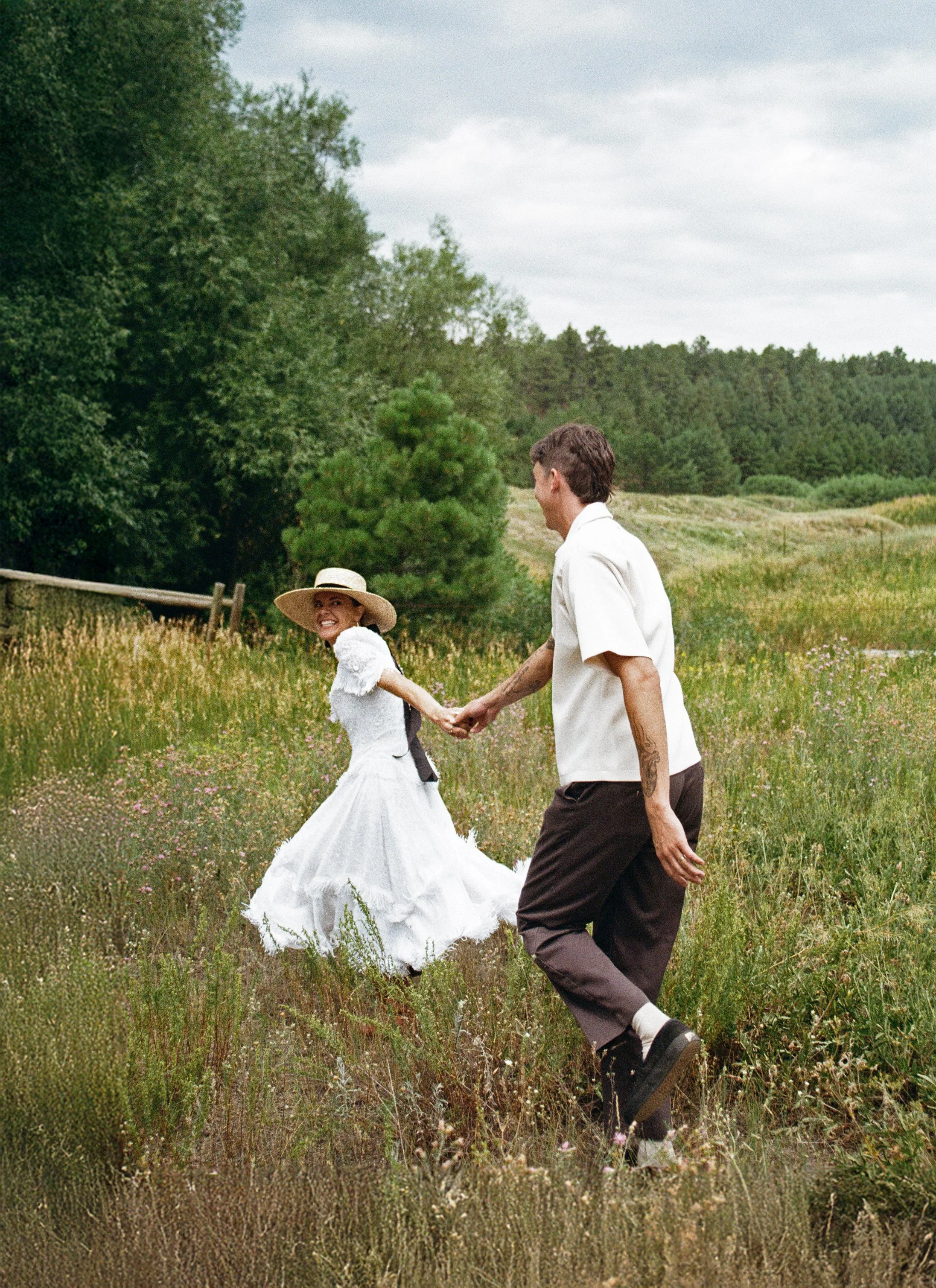 A couple holding hands and dancing in a field with tall grass and wildflowers, with trees and a cloudy sky in the background.