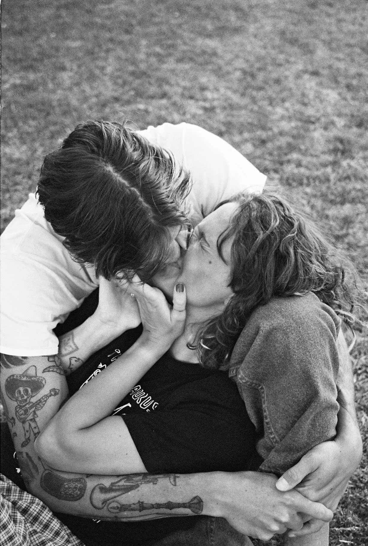 A black and white photo of two women kissing, one with long curly hair and the other with short dark hair, outdoors on grass.
