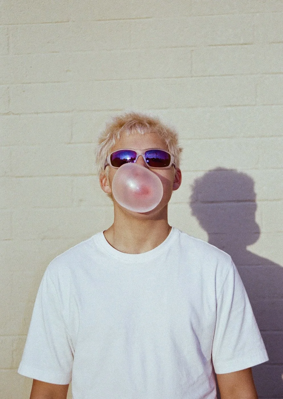 Young person with blonde hair wearing sunglasses blowing a pink bubble from gum, standing against a beige brick wall, casting a shadow.