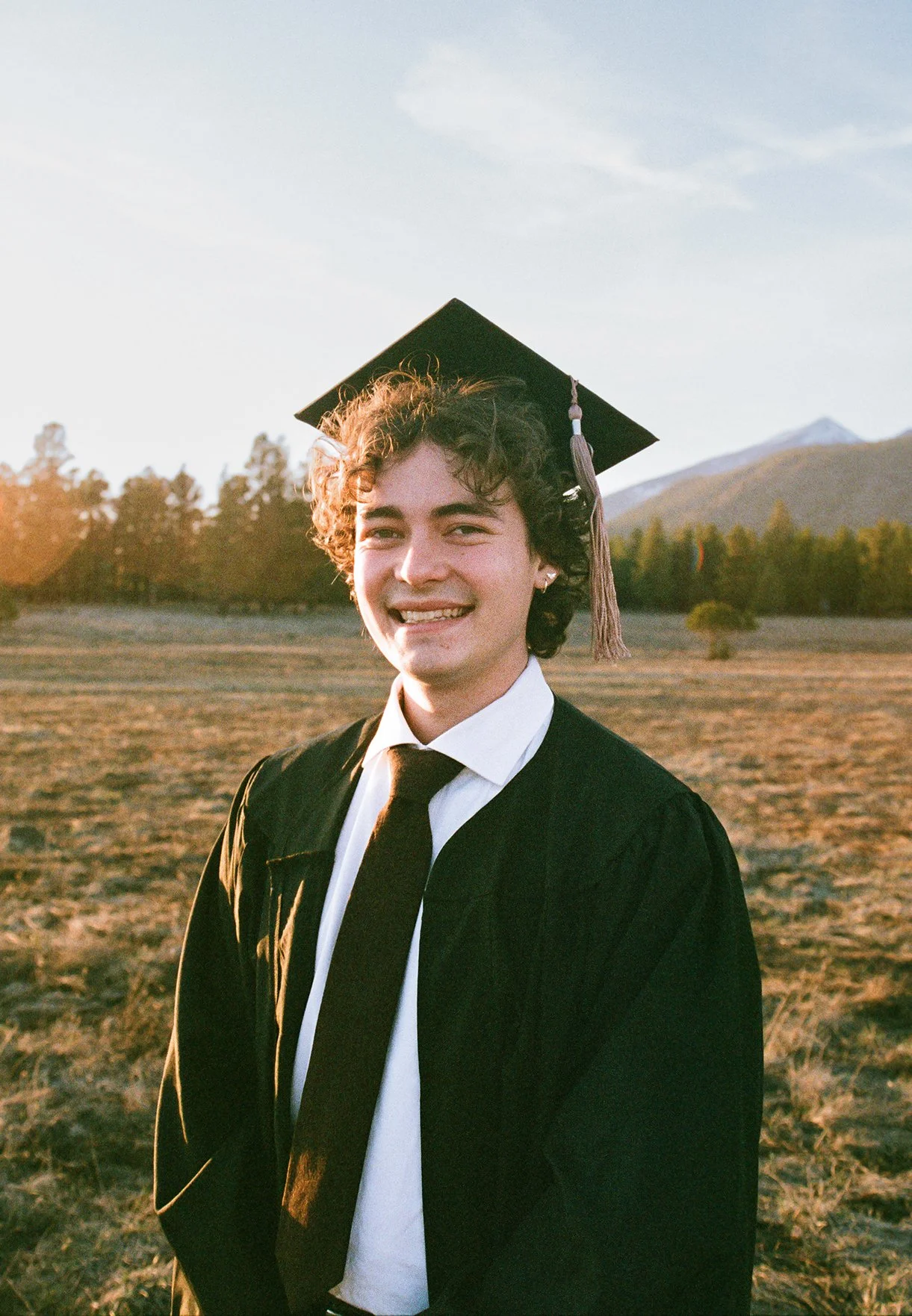 A young person in graduation attire, wearing a black cap and gown, smiling outside during sunset with mountains and trees in the background.