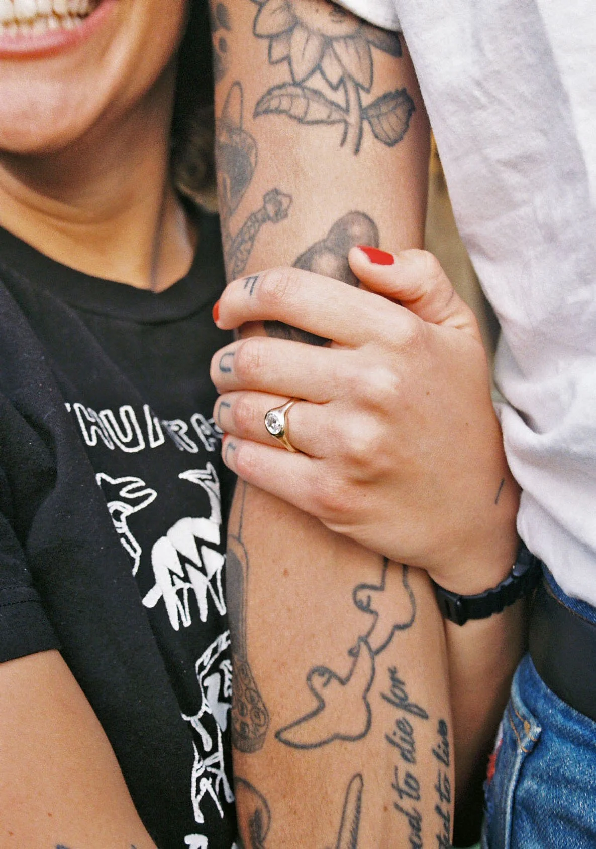 Close-up of two people hugging, showing tattoos on their arms. One person has red painted nails, a ring with a large gemstone, and tattoos including a flower, a sword, and a quote. The other person is wearing a black T-shirt with white graphics.