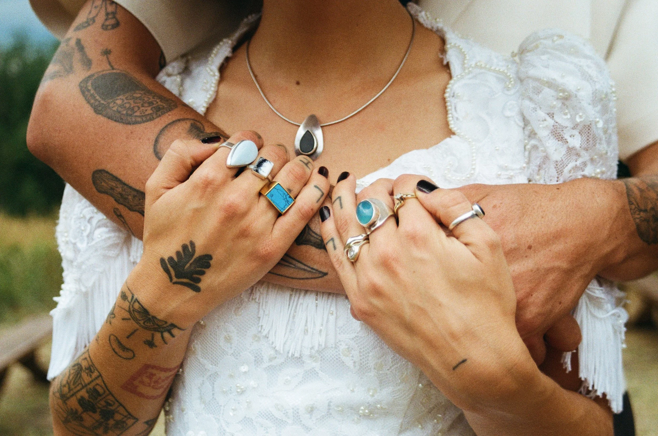 Close-up of two women holding hands with their fingers intertwined, wearing multiple rings and jewelry, with tattoos visible on their arms and hands, against a background of a white lace dress.