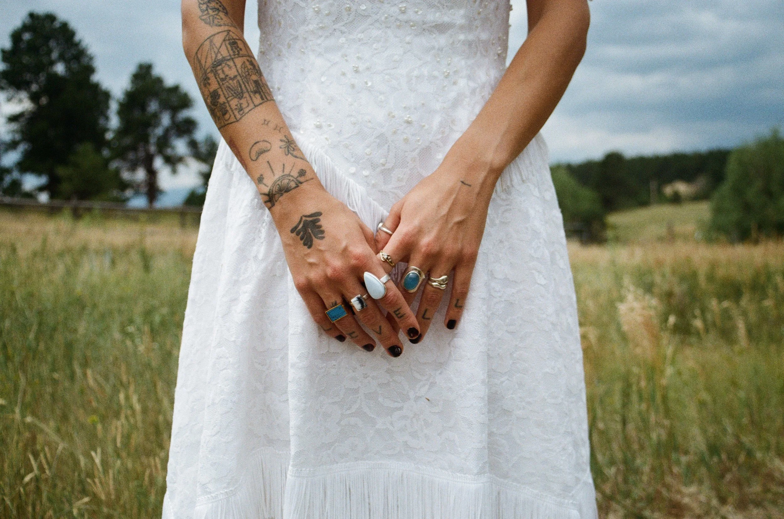 Person wearing a white lace dress, showing tattoos on their arms and fingers, standing in a grassy field with trees in the background.