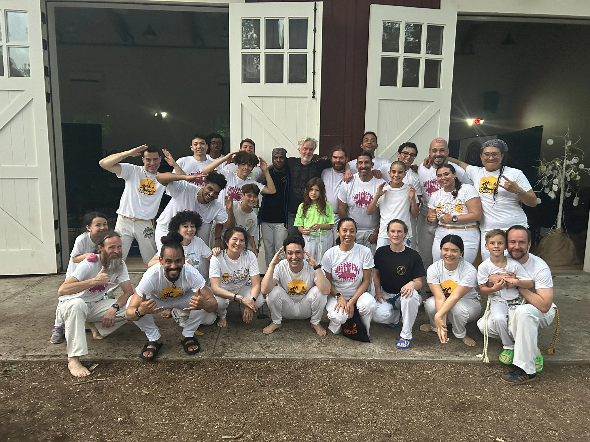 Group of people posing together outside, in front of a large barn door in Cold Spring NY, some doing capoeira poses, dressed in capoeira uniforms and casual clothing, after a gathering for their graduation event.