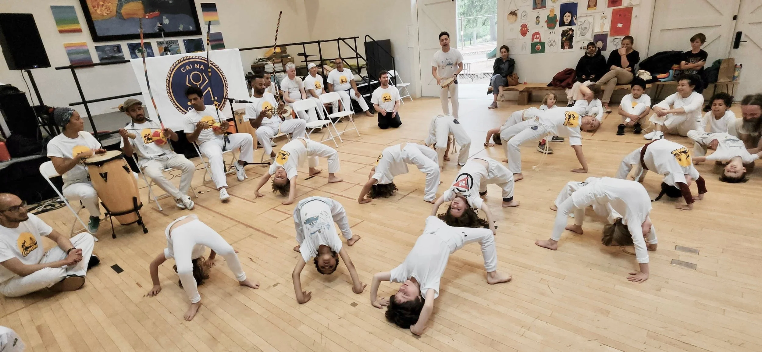 Children and adults in white capoeira uniforms practicing capoeira, some doing backbends, in a large room with wooden floors. Several seated capoeira teachers play percussion instruments, and some audience members sit against the wall.