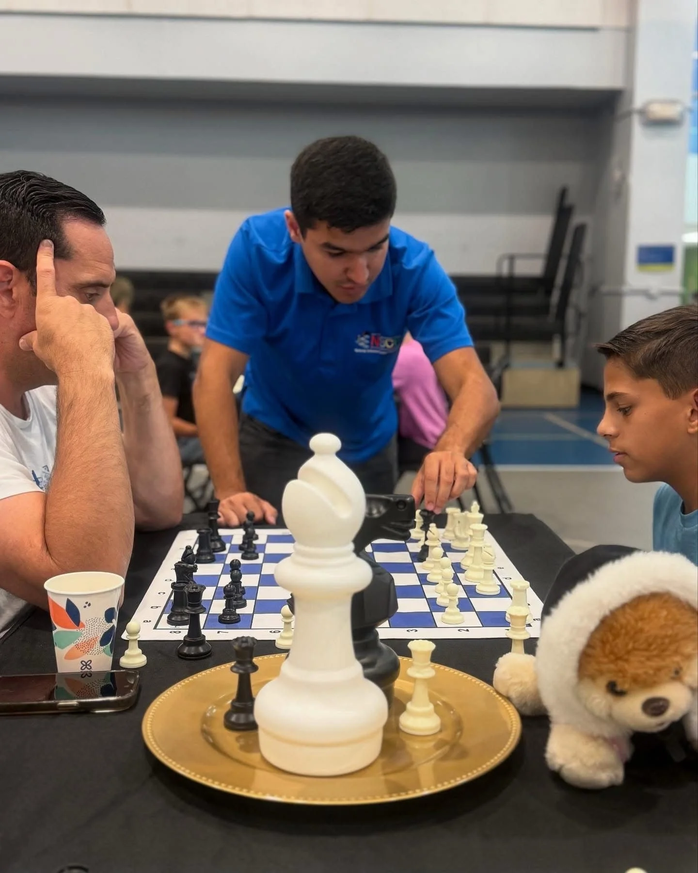 People playing chess at a table with a stuffed dog and a large decorative chess piece in the foreground.
