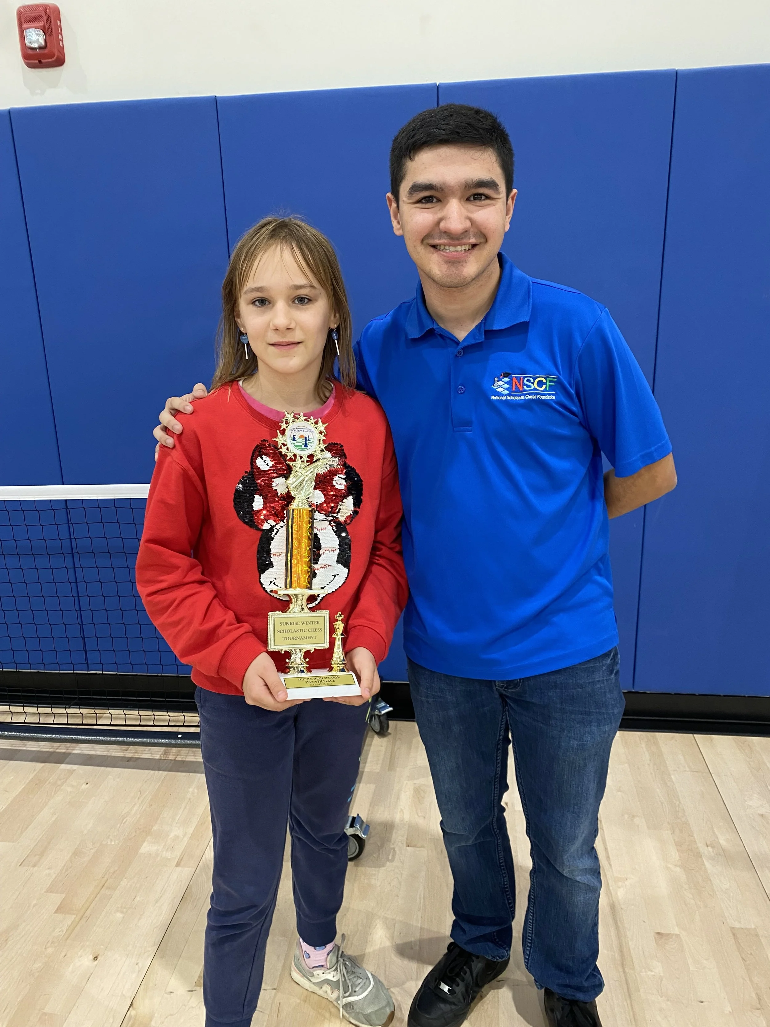 A young girl and an adult man standing in a gym, smiling at the camera. The girl is holding a trophy and wearing a red sweatshirt with a ladybug design, and the man is wearing a blue polo shirt with a logo. The background has blue padding and a gym net.