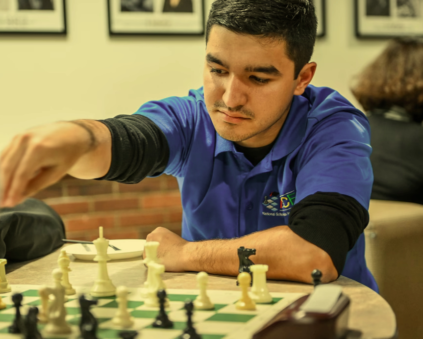 Young man playing chess at a table in an indoor setting with framed pictures on the wall.