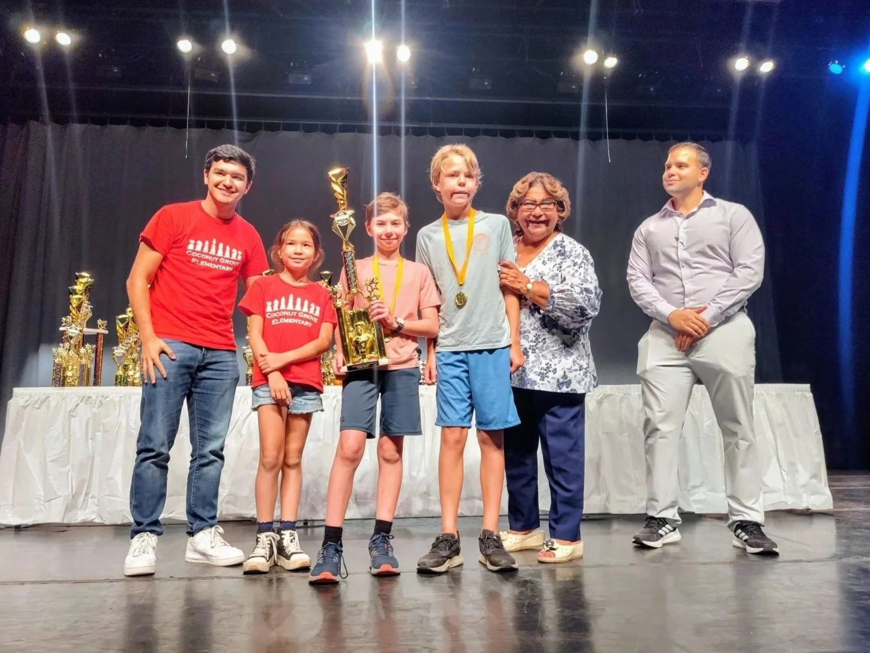 Group of children and adults on stage at an award ceremony, with children holding a trophy and medals, smiling and posing for a photo.