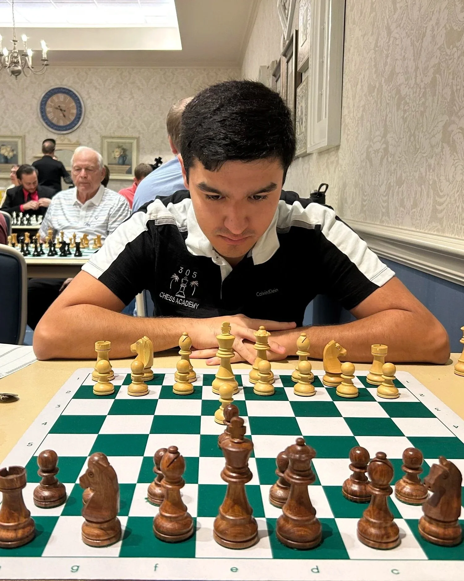 A young man playing chess in a room with wallpaper and framed pictures on the wall, with other people in the background also engaged in chess games.