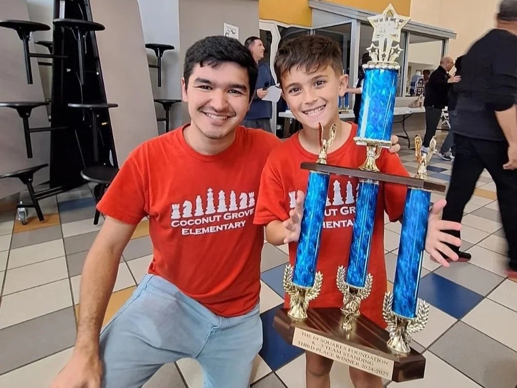 Two boys in red shirts are smiling and holding a large blue trophy with four blue columns and gold trophies, at an indoor event.