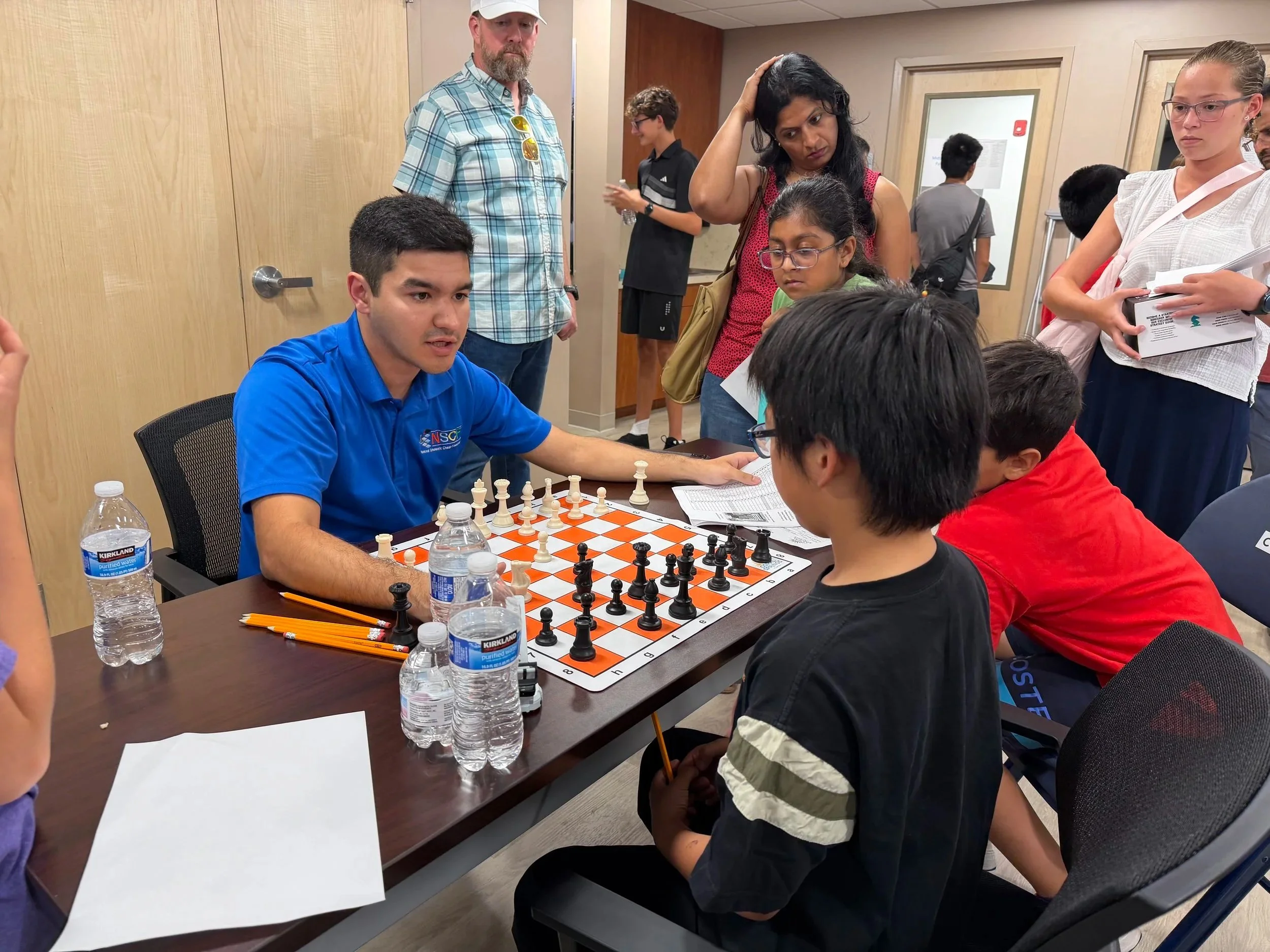 People playing chess at a meeting or tournament, with spectators watching, in a room with wood-paneled walls.