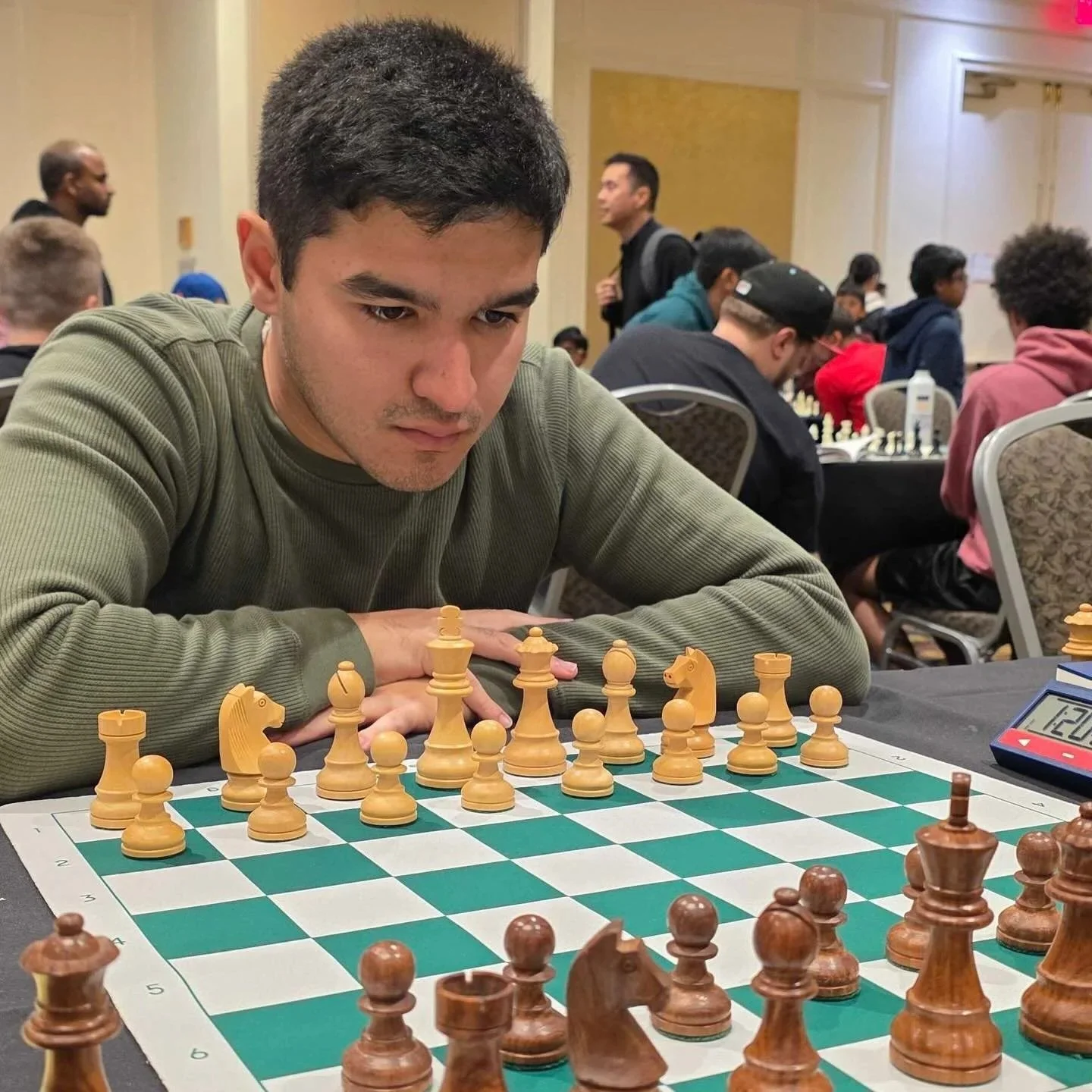 A young man with dark hair and a green sweater concentrating on a chess game at a tournament, with other players and spectators in the background.