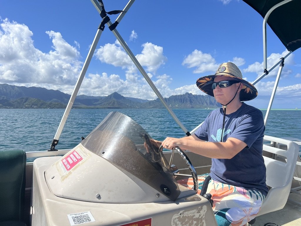 A man wearing sunglasses, a wide-brimmed hat, a blue t-shirt, and colorful shorts is steering a boat on a body of water with mountains in the background. The sky is blue with scattered clouds. considering adoption for her child and learning how to ge