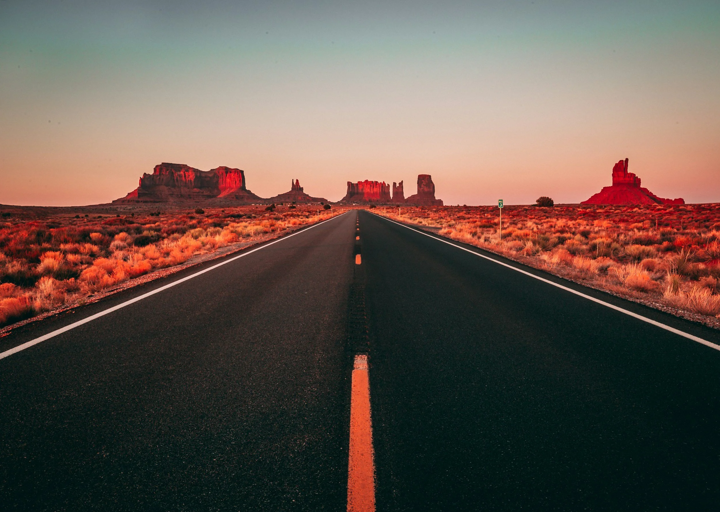 A straight open road in a desert landscape with rock formations at sunset.