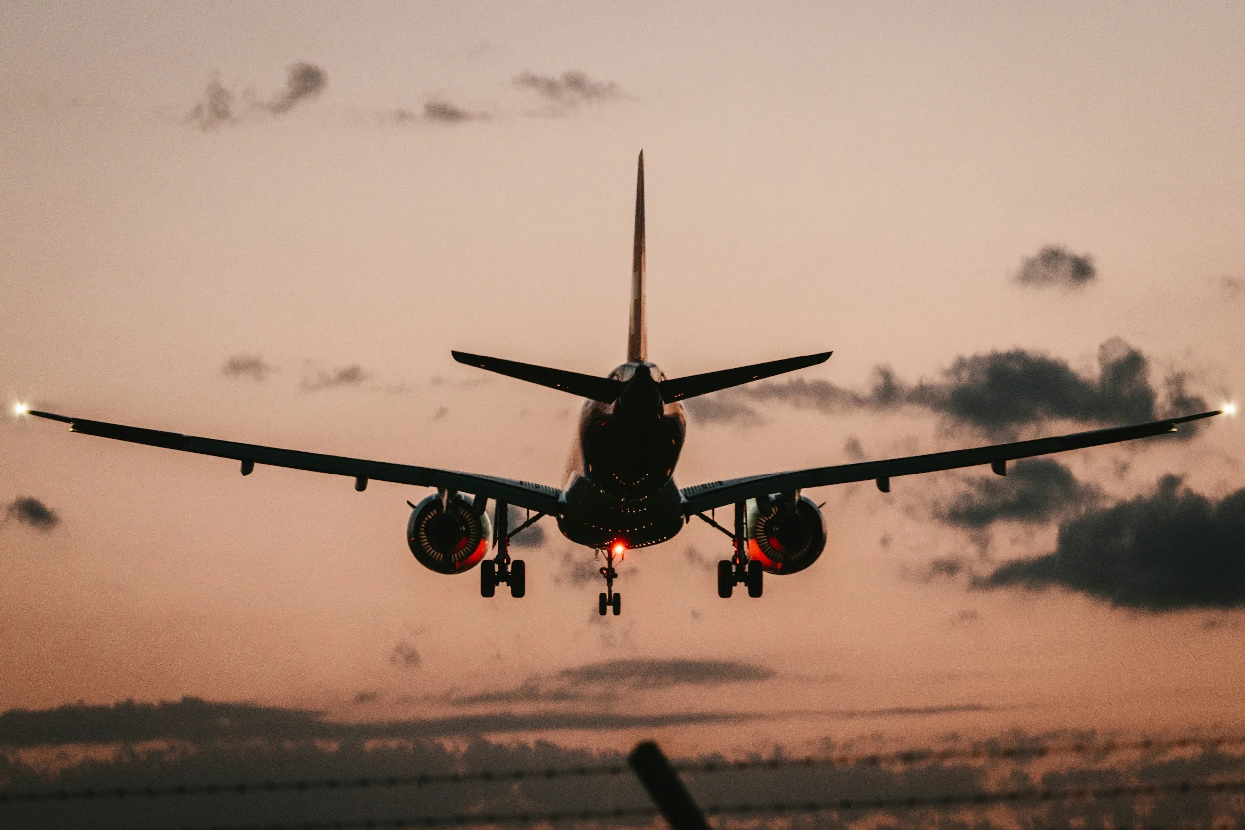 An airplane approaching for landing during sunset with a pinkish sky, clouds, and silhouettes of mountains in the background.
