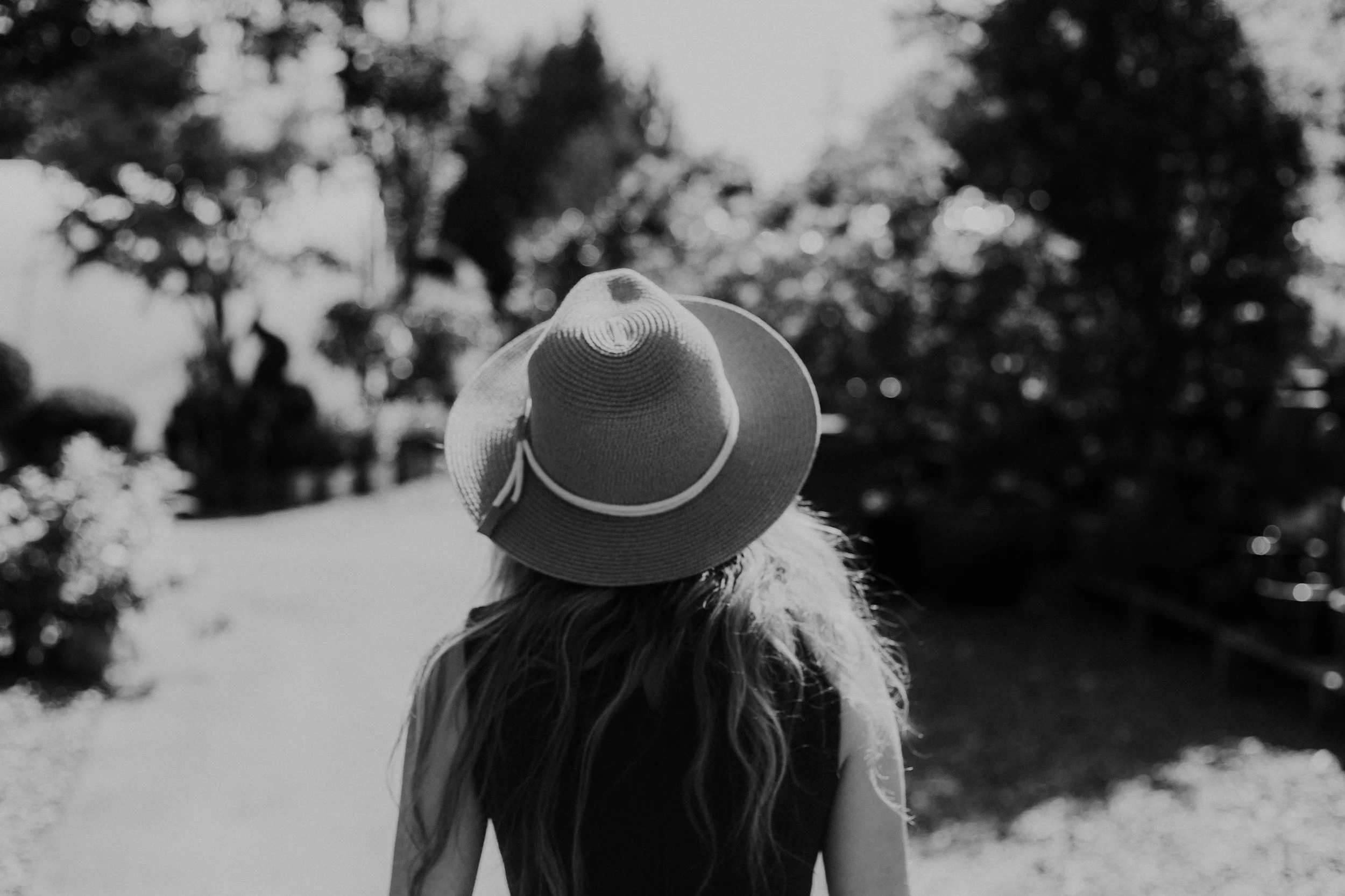 A woman with long hair wearing a wide-brimmed hat walking outdoors on a sunny day, with trees in the background.