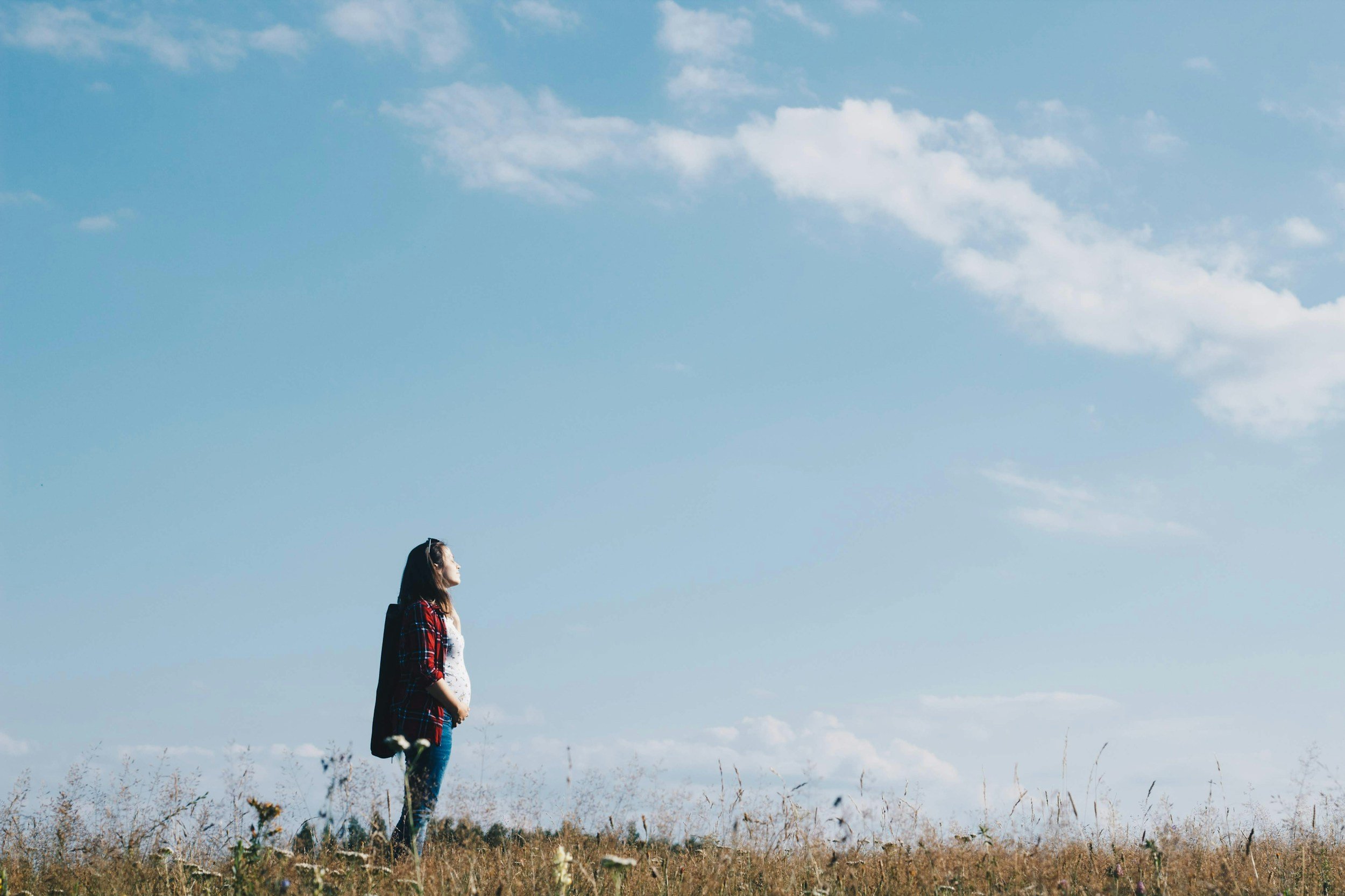 A woman standing in a grassy field under a blue sky with scattered clouds, facing to the left.