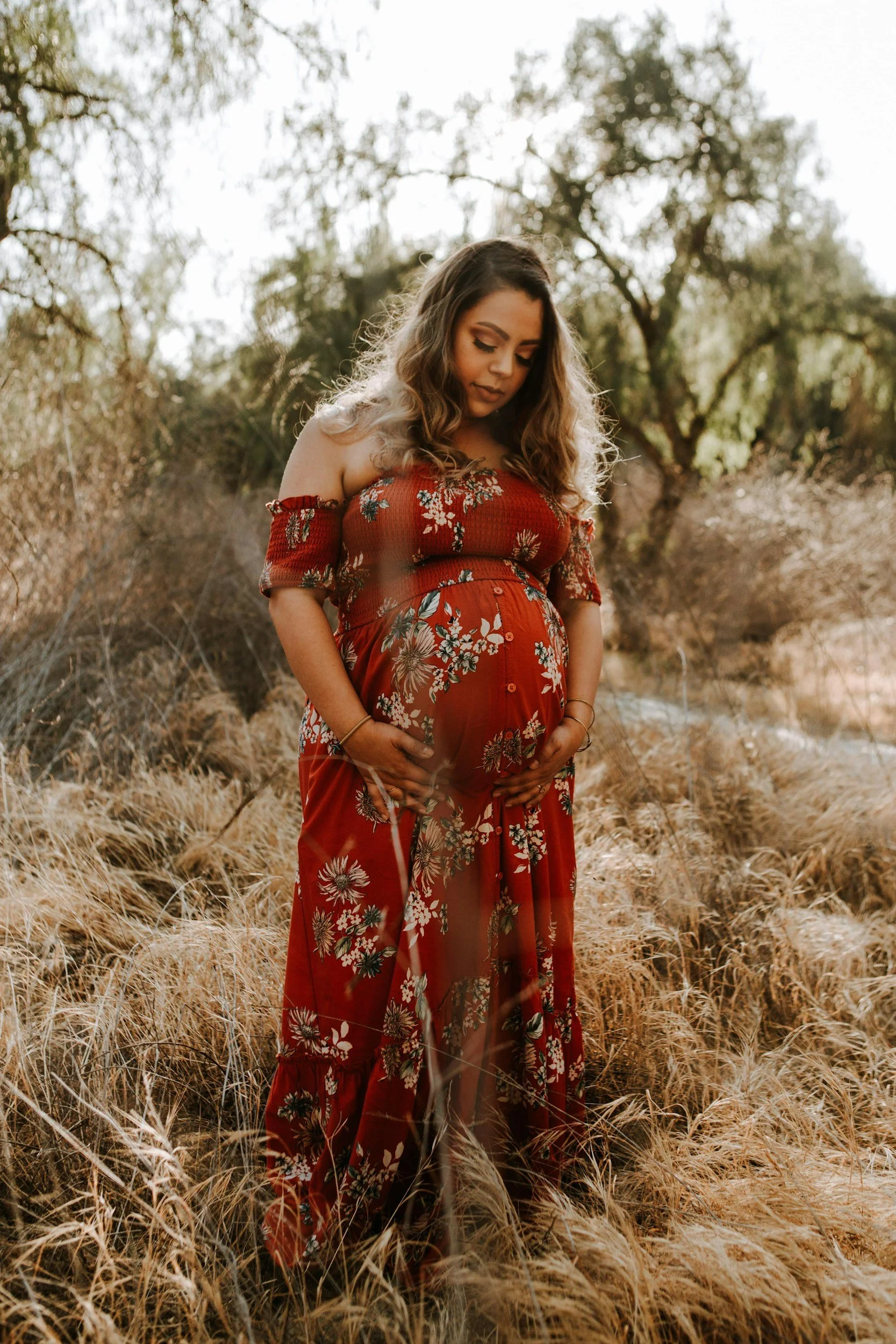 A pregnant woman dressed in a red floral off-the-shoulder dress stands in a field of dry grass, touching her belly and looking down.