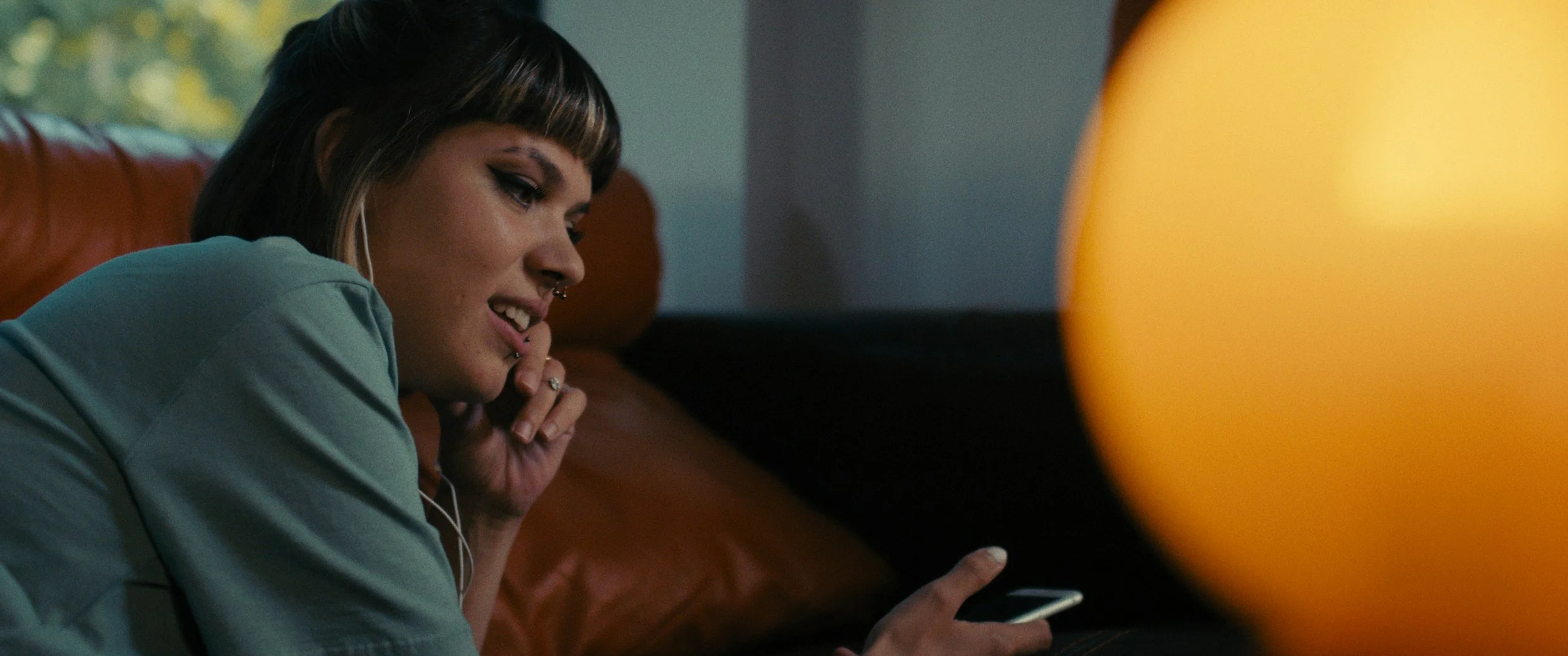 A young woman with dark hair and bangs, wearing a light-colored shirt, is lying on a brown leather couch in a dimly lit room, looking at her phone and smiling considering adoption for her child and learning how to get pregnant help options to create
