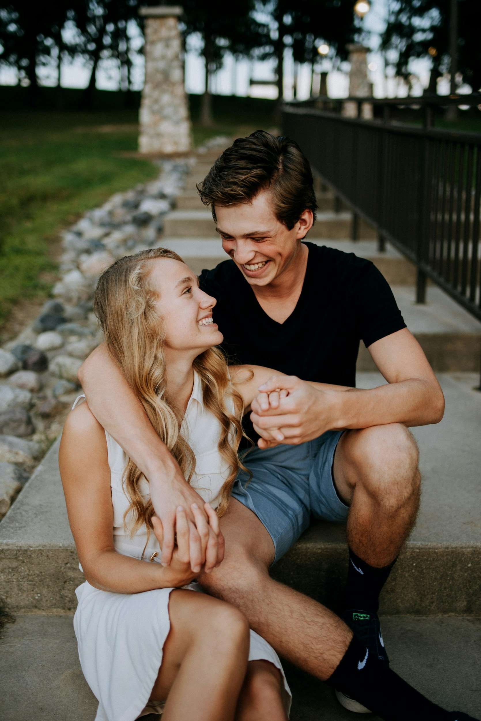 A young man and woman sitting on outdoor steps, smiling and looking at each other lovingly, with trees and a black railing in the background. considering adoption for her child and learning how to get pregnant help options to create an adoption plan.