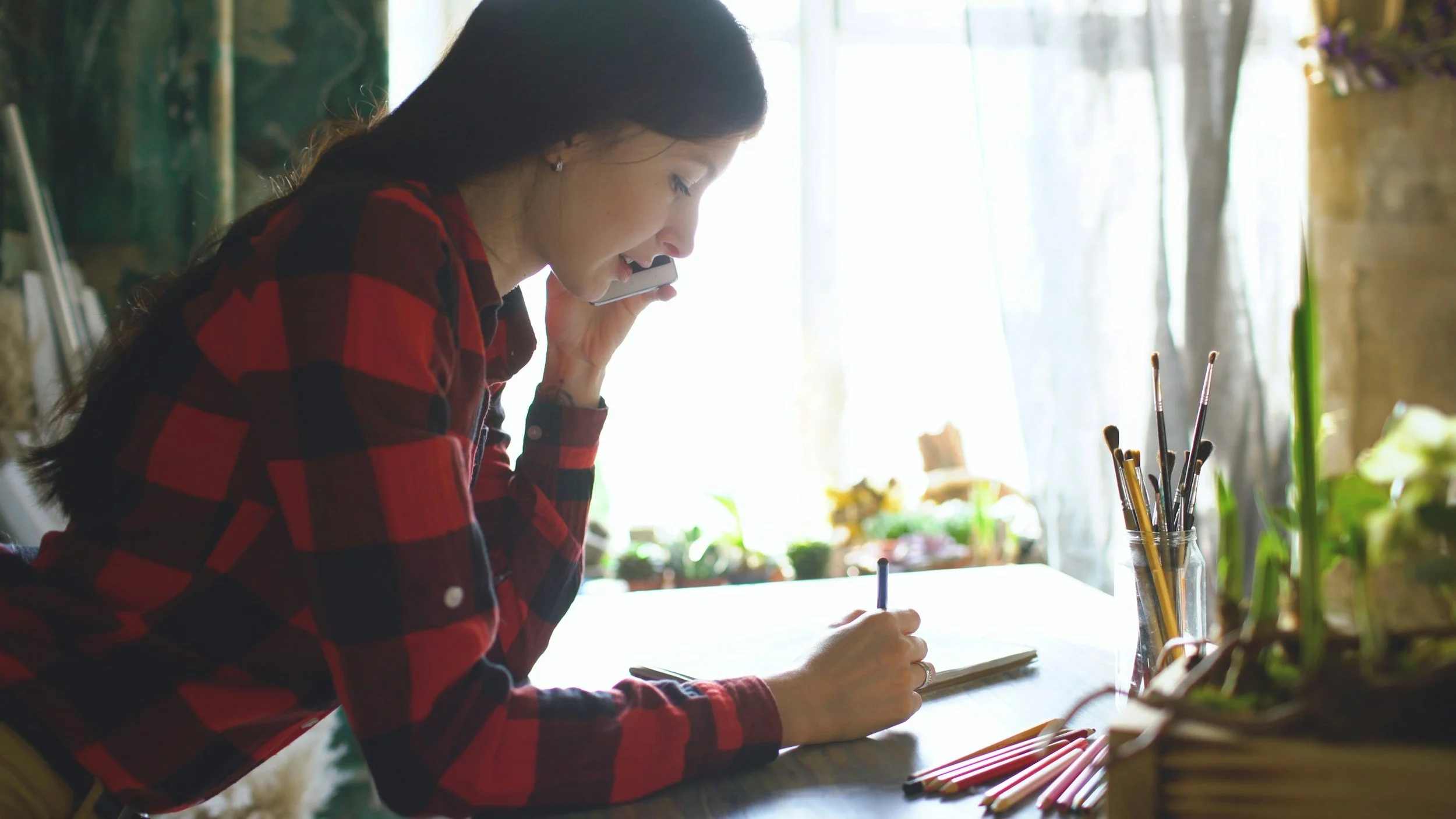 A woman in a red and black plaid shirt sitting at a table, talking on her cellphone, writing in a notebook, surrounded by art supplies and plants in a brightly lit room.