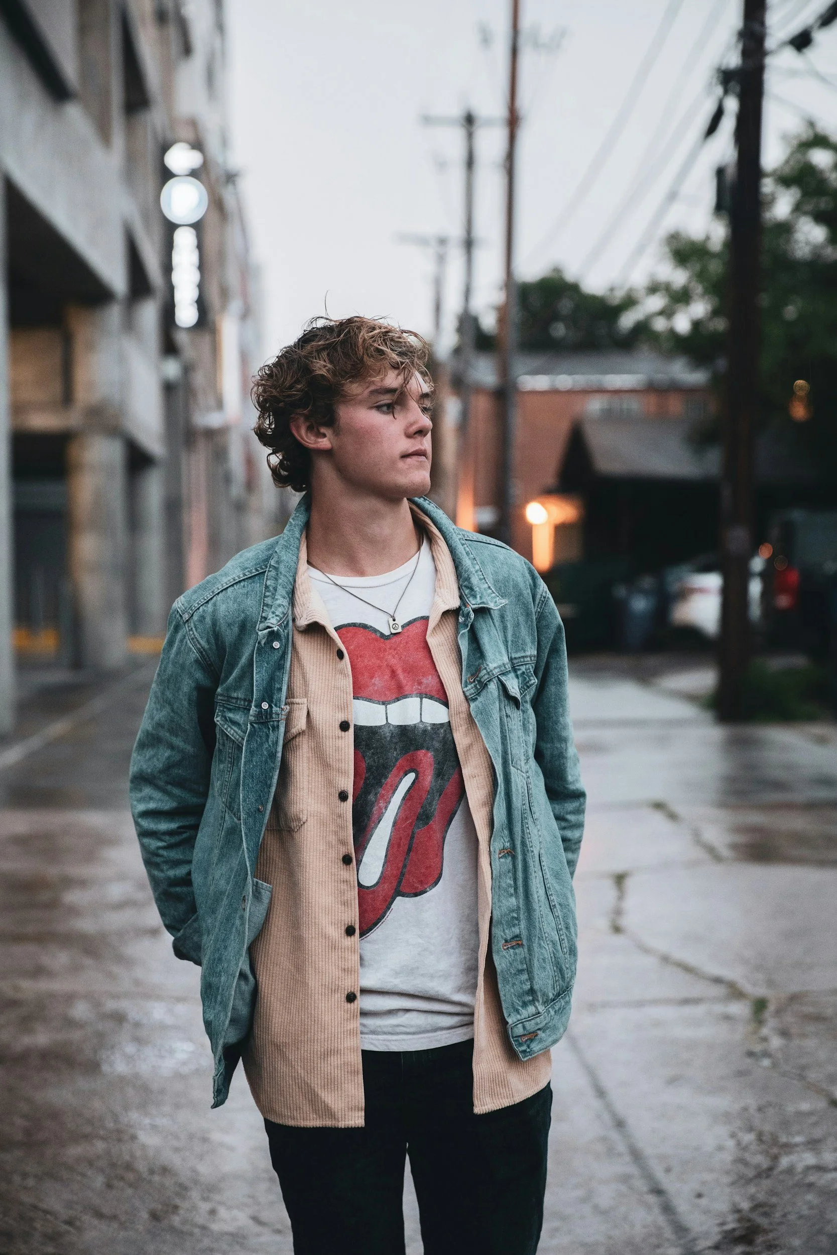A young person walking down an alleyway during the evening or late afternoon, wearing a denim jacket over a graphic t-shirt and dark pants, with their hands in their pockets and gazing to their right.