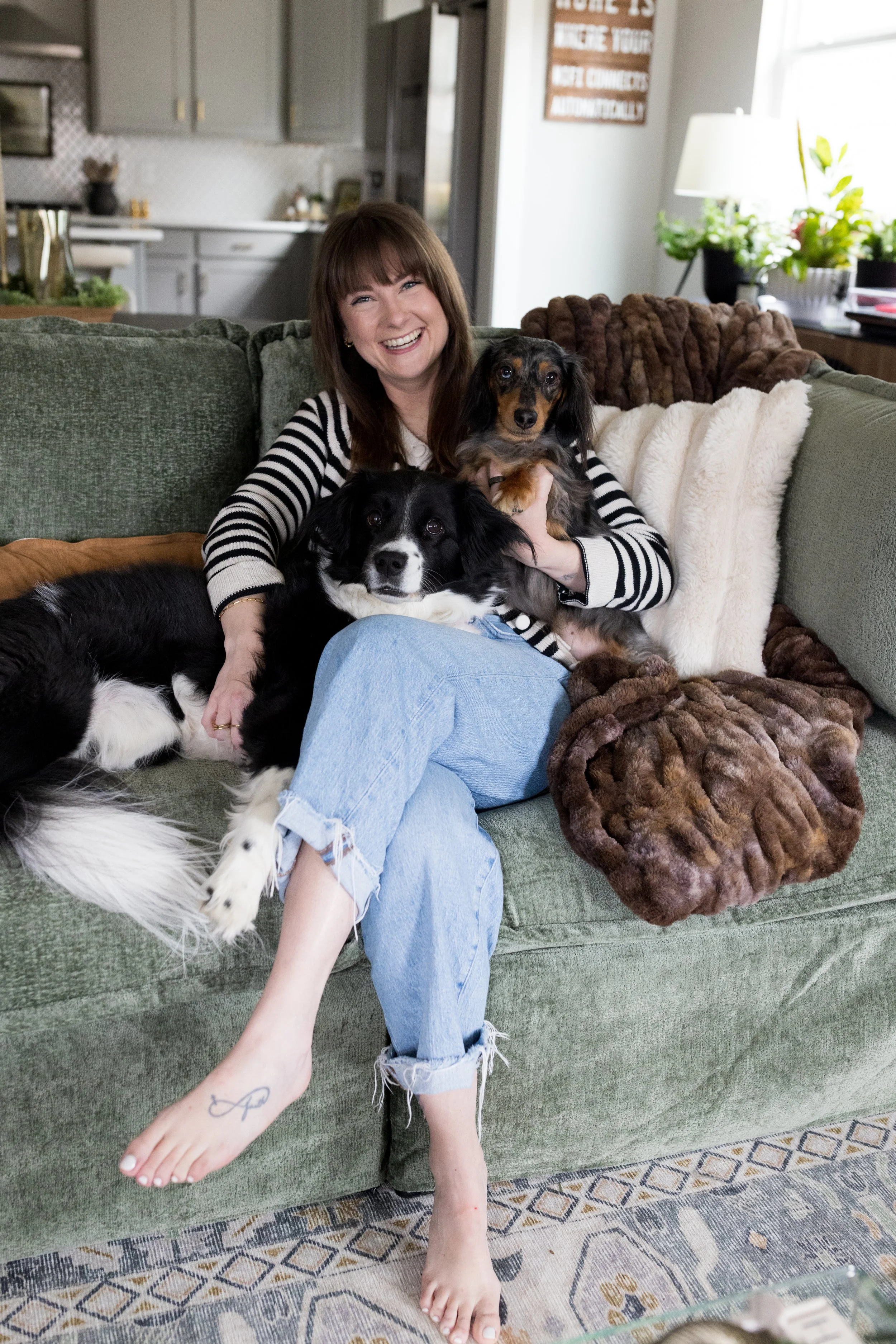 A woman with shoulder-length brown hair, wearing a black and white striped shirt and light blue ripped jeans, sitting on a green sofa, smiling, with two dogs. One dog is a black and white Border Collie resting on her lap, and the other is a small, long-haired Dachshund with a dark coat with tan markings, held in her arms. The sofa is decorated with a fur blanket and pillows. The background shows a living room with a plant, a lamp, and a sign on the wall.