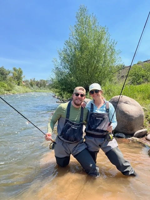 A man and woman fishing in a river, standing in shallow water with fishing rods, wearing waders and sunglasses, with trees and rocks in the background on a bright, sunny day.