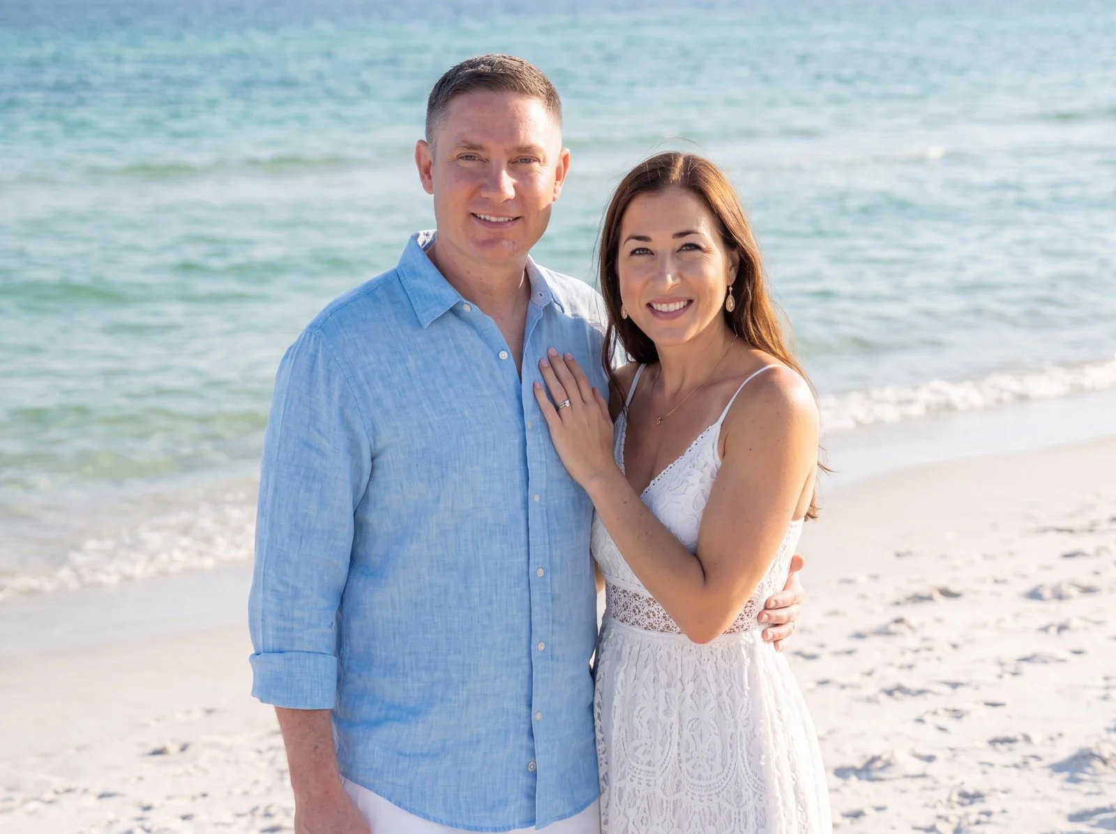 A smiling couple standing on the beach near the ocean, with the woman showing her hand with a ring on her finger. As they contemplate adopting an infant child through private adoption. with Starr Adoption Services